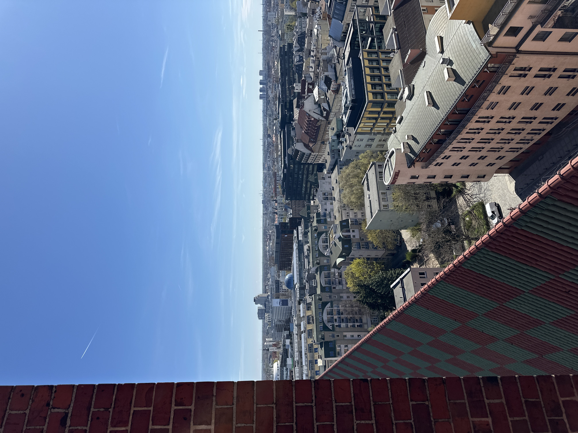 Here is a detailed description of the image:
The image presents a high-angle, long-range view of a cityscape, possibly taken from a tall building or tower. The scene includes a clear blue sky dominating the left side of the frame, indicating daytime. Contrails from aircraft are visible in the sky.
The foreground features architectural elements, including a brick wall and a red and green checkered tile roof, suggesting that the photo was taken from an elevated position, like a rooftop or tower.
In the mid-ground, a dense urban landscape extends into the distance. Numerous buildings of varying heights and architectural styles can be seen, characterized by flat roofs and rectangular facades. Some buildings have visible windows and architectural details, such as rooftop structures. Trees and green spaces are interspersed among the buildings, suggesting parks or residential areas.
The perspective of the photo provides a sense of depth, with the city extending towards the horizon where the cityscape merges with the sky. The overall impression is that the photograph captures a sprawling European city on a clear day.
While the specific buildings and landmarks that would confirm the location are not discernible without further context, the architectural styles and urban density are consistent with the city of Wrocław in Poland. However, further clues or landmarks would be needed to confirm the precise location.