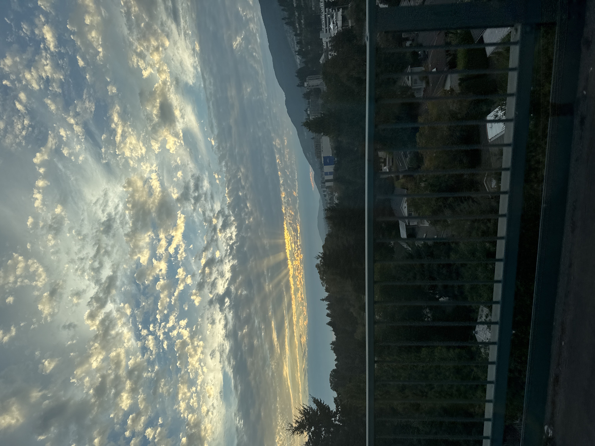The image captures a view from a bridge in West Vancouver, Canada, overlooking a residential area with a backdrop of forested hills and mountains. The sky is filled with a dramatic cloudscape, with rays of sunlight breaking through at the horizon, suggesting either sunrise or sunset. In the foreground, a dark green metal railing runs horizontally across the bottom of the frame, obscuring a portion of the view. Below the railing, houses and trees are visible, nestled amongst the greenery. Further in the distance, buildings and more trees dot the landscape, leading up to the imposing, tree-covered mountains. The overall impression is one of a serene, natural setting within an urban environment.