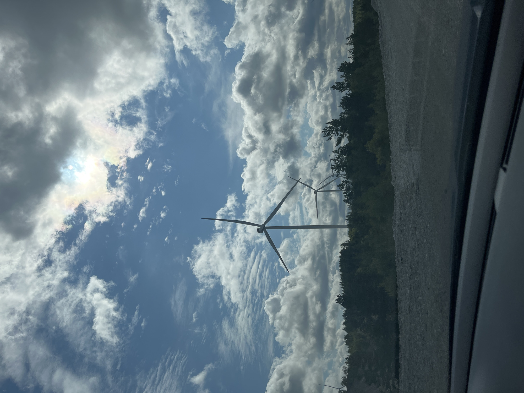 The image shows a view of a wind farm in Aidu, Estonia. Several wind turbines are visible in the middle ground, silhouetted against a bright, cloudy sky. The turbines have tall, white towers and large, dark blades. In the foreground, there is a rough, gravelly surface, likely a road or path. Behind the turbines, a dense line of dark green trees forms a forest. The sky is partly cloudy with patches of blue visible. The sun is partially obscured by clouds, creating a bright, slightly hazy effect with some iridescent colors visible in the clouds near the sun. The overall impression is of a vast, open landscape dominated by renewable energy infrastructure.