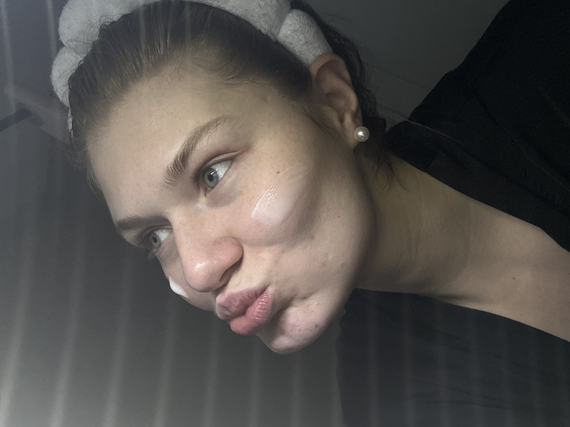 A young adult female is depicted in a close-up, upward-angled shot. Her hair is secured back from her face with a white, plush headband. She wears a dark garment, visible on her left shoulder and chest. A single spherical pearl earring is present on her right earlobe. Her lips are pursed forward in a "kissing" expression, and her light-colored eyes are directed upwards and slightly left. Three distinct applications of a white or translucent cream/lotion are visible on her face: one above her right eyebrow, one on her right cheekbone, and one on the left philtrum area above her upper lip. Her skin shows some minor texture and scattered small blemishes. The background is a solid, dark gray, suggesting an indoor setting with diffused lighting from the upper left. The scene is observed in Kazan, Russia.