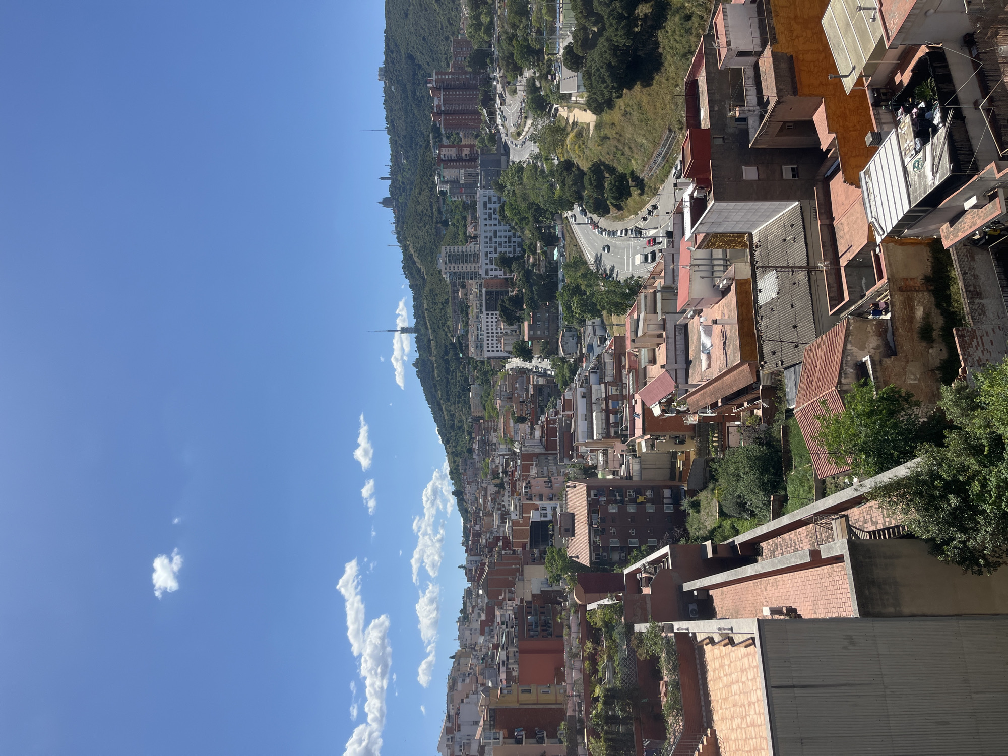 The image shows an elevated view of a section of Barcelona, Spain, characterized by densely packed buildings with red-tiled roofs and various architectural styles. To the left, a hillside covered in trees and buildings rises, with roads winding along its contour. A clear blue sky with scattered cumulus clouds is visible above. In the foreground, rooftops of neighboring buildings provide additional context. The overall scene conveys a sense of urban density and the city's integration with the surrounding natural landscape.