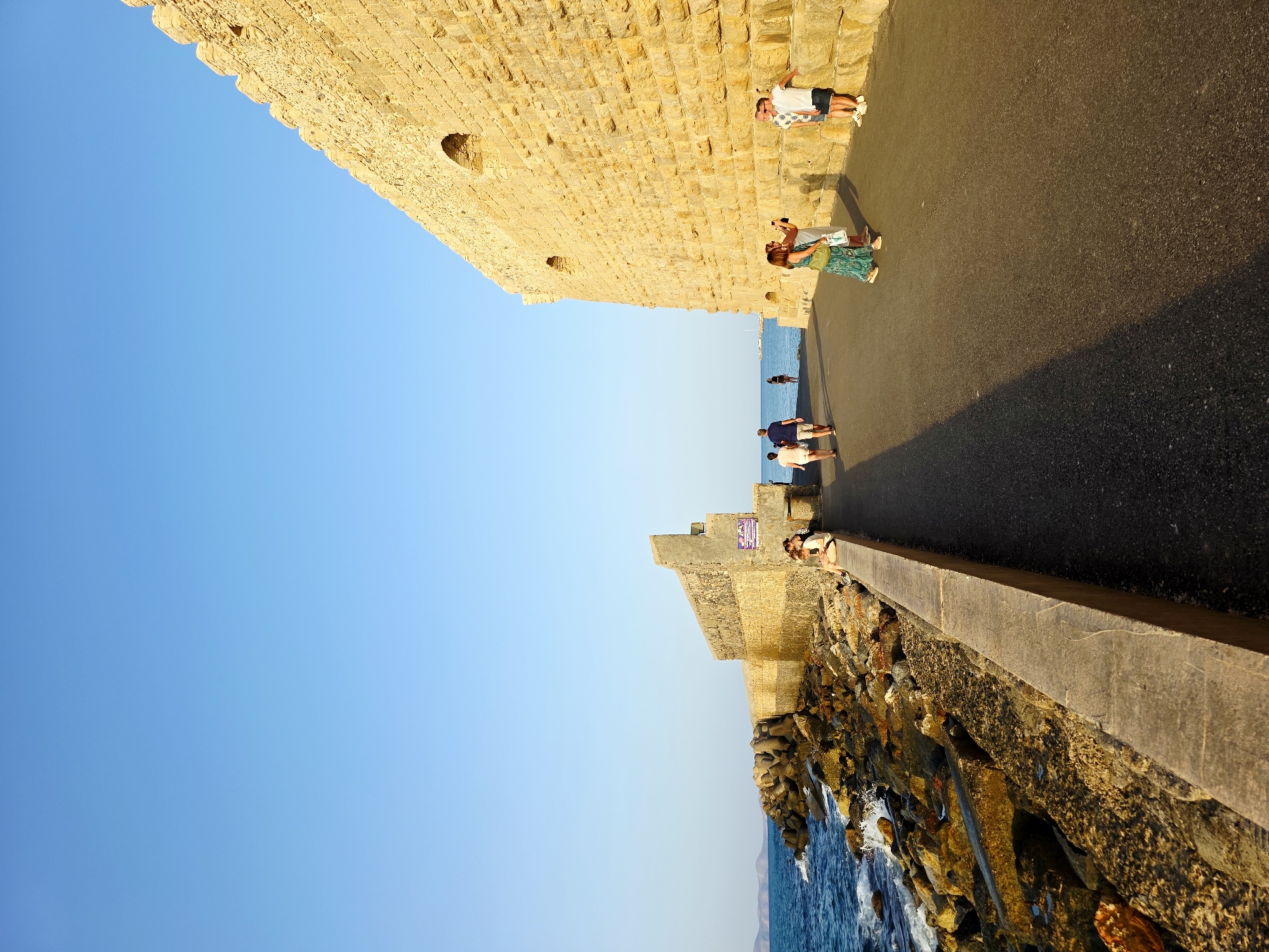 This is a landscape photograph taken at the Venetian harbor in Heraklion, Greece. The image depicts a wide, paved walkway running alongside a massive, ancient stone wall. The wall, constructed from large, rough-hewn yellowish stones, dominates the right side of the frame and exhibits a weathered, robust appearance with several arched openings visible. The walkway is dark asphalt and slopes gently downward towards the left.
To the left of the walkway, a rocky breakwater extends into the blue sea, with large, angular concrete tetrapods and natural boulders forming a protective barrier against the waves. The sea is a vibrant blue with whitecaps indicating moderate wave activity. The sky above is a clear, bright, and cloudless cerulean blue.
Several people are present in the scene. In the foreground on the right, two individuals are interacting near the stone wall; one appears to be taking a photograph of the other with a mobile phone. Further along the walkway, a group of three people are walking away from the camera, and another individual is seated on the low concrete barrier of the breakwater. In the distance, more people are visible walking along the pathway and near the breakwater. The sun is low in the sky, casting long shadows across the walkway, suggesting it is either late afternoon or early morning. The overall atmosphere is one of historical grandeur meeting a pleasant coastal environment.