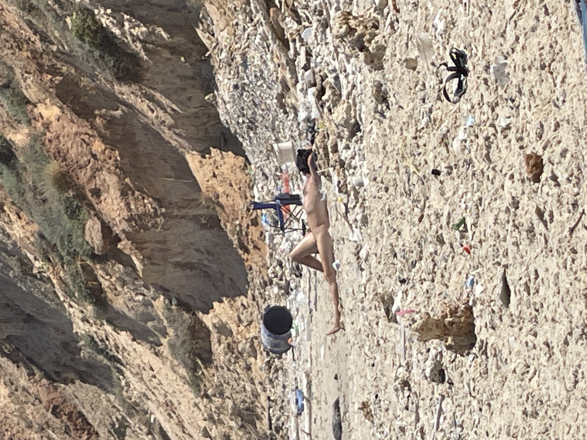 The image depicts a rocky, debris-strewn beach area near the cliffs of Ga'ash, Israel. In the foreground, a person is lying on their back on the sand, with their legs extended. They are unclothed. The beach is littered with various objects, including a large black barrel to the left of the person, a blue cooler, a collapsed camping chair, and piles of what appears to be rubble and trash. The background shows a steep, eroded cliff face with sparse vegetation. The overall scene suggests a neglected and possibly polluted coastal environment.