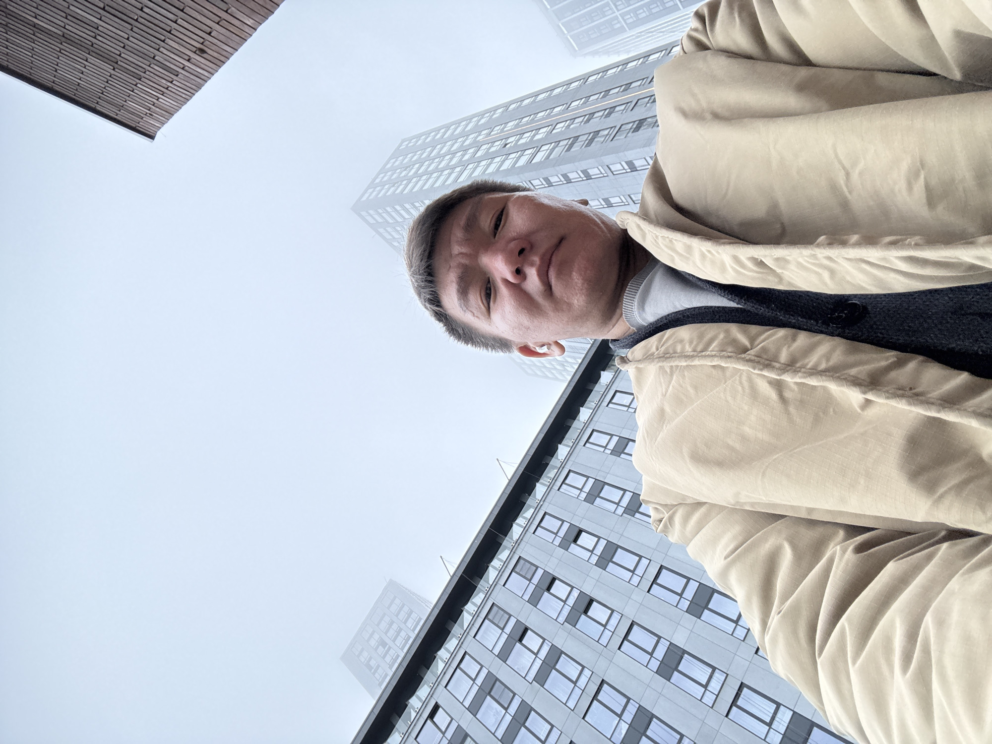 The image shows a man looking down at the camera while standing in an urban environment. He is wearing a tan jacket over a grey sweater. Several tall buildings surround him, some partially obscured by fog. One building has a grid pattern of windows. Another building has a wooden design on part of it. The buildings suggest a city location, specifically Moscow, Russia, based on the architectural style. The sky is overcast. The scene is shot from a low angle, emphasizing the height of the buildings and the man's position looking downward.