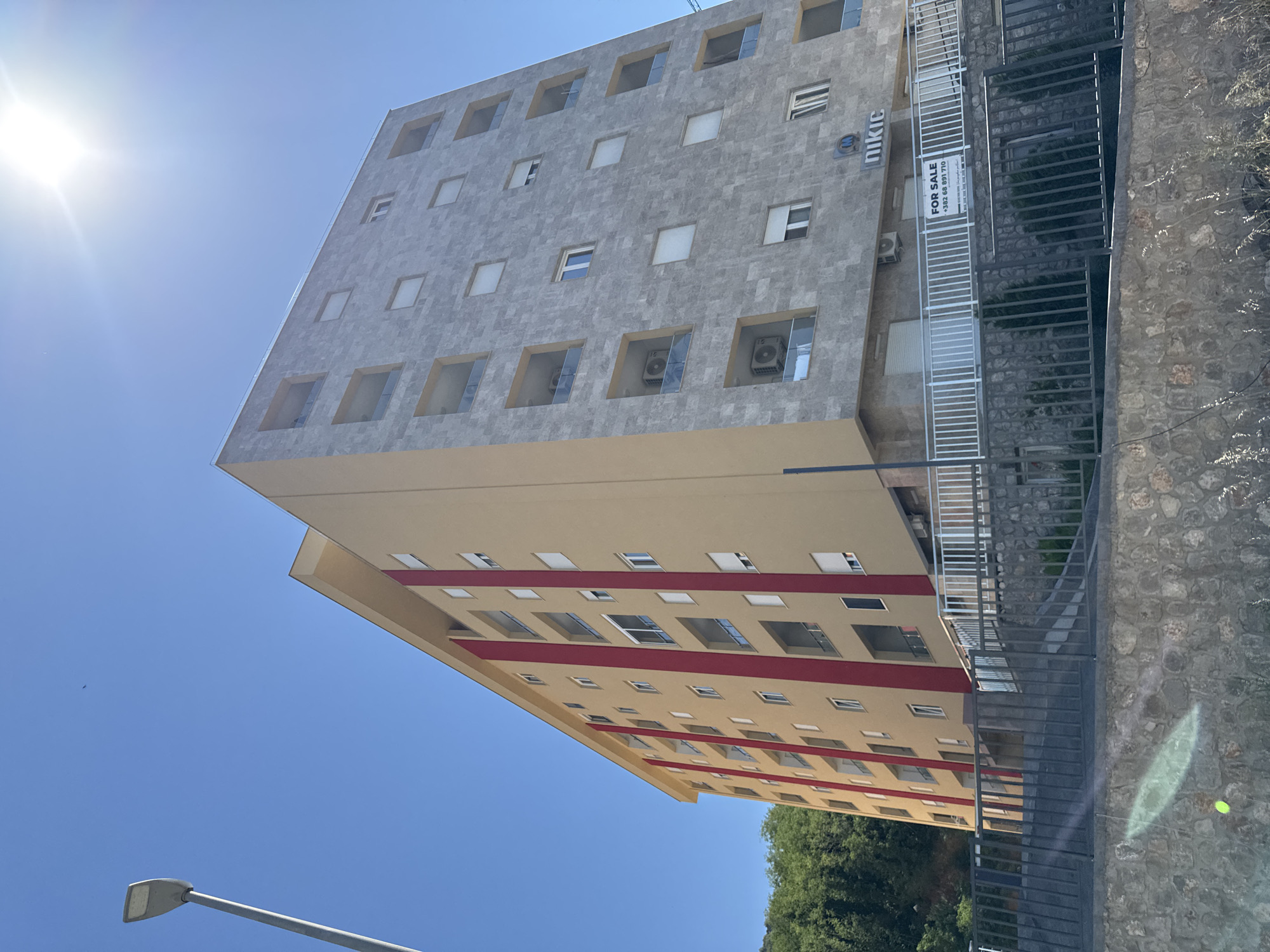 An eye-level, medium shot captures a multi-story building in Boreti, Montenegro, under a clear, sunny sky. The building is divided vertically, with the left half painted a warm beige and featuring three prominent red vertical stripes. The right half of the building is clad in light gray stone tiles, with numerous square windows and balconies. Some windows appear to have white blinds, and air conditioning units are visible on some balconies. A sign that reads "FOR SALE +382 68 891 710" is affixed to a railing in front of the building's ground level. A streetlamp stands to the left of the building, and a lush green hillside with trees is visible behind the left side. The foreground consists of a stone wall and a metal fence. The sun is bright, creating a lens flare in the upper right quadrant of the image.