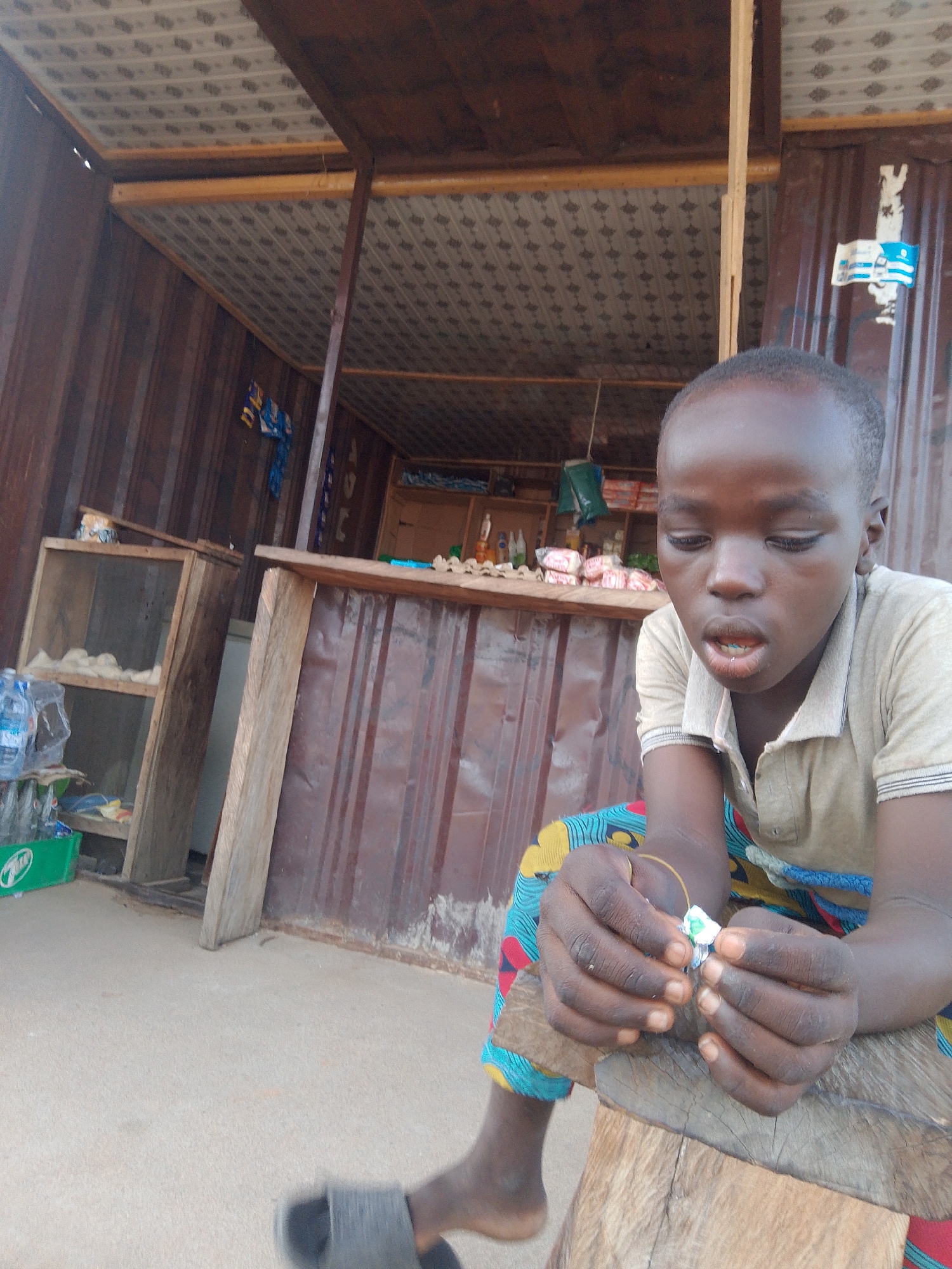 A young child is sitting on a wooden stool in front of a small stall. The stall appears to be made of corrugated metal and wood, with shelves displaying various items, including what seem to be packaged goods like soap or snacks, and bottles of water. The child is wearing a light-colored polo shirt and brightly patterned pants. Their hands are clasped, and they are looking down at something small they are holding, perhaps a bead or a piece of candy, with a focused expression. The stall's roof is covered with a patterned material, and wooden beams support the structure. In the background, more shelves are visible, and a small flag is attached to the corrugated wall. The ground is a light-colored, sandy surface. The lighting suggests it might be daytime, possibly late afternoon, given the soft shadows. The overall scene depicts a casual moment at a roadside market stall.