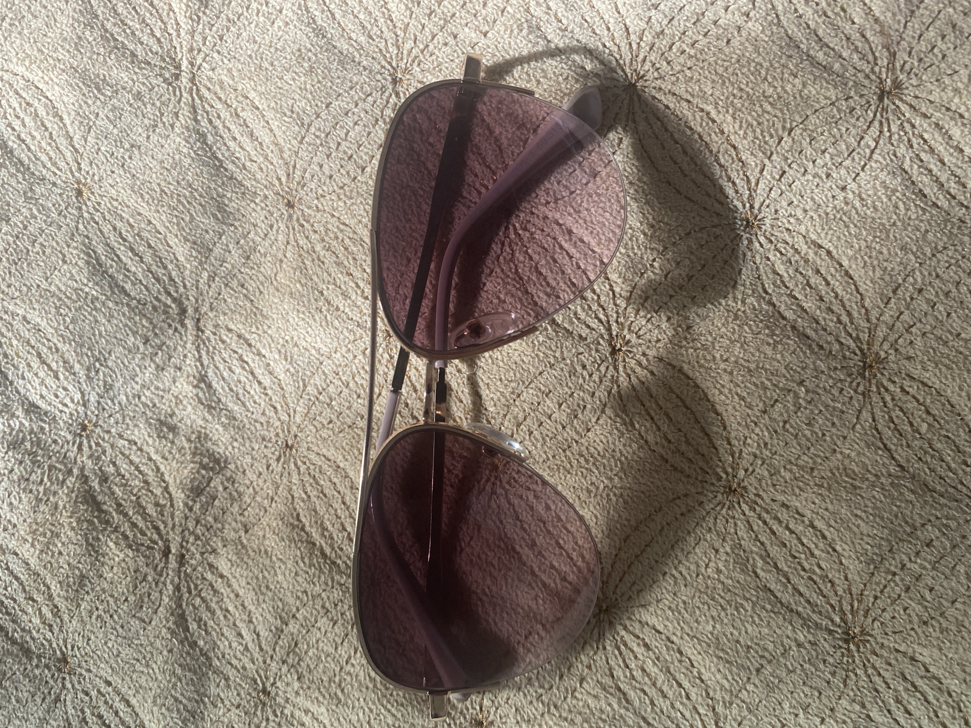 A pair of aviator sunglasses with gradient pink lenses rests on a textured, light-colored fabric with a repeating circular, floral-like pattern. The sunglasses have thin gold-toned frames and pink arms. Shadows from the sunglasses and an unseen object are cast across the fabric, indicating a light source from the upper left. The scene is a close-up, overhead shot. There is no discernible information about the location in Kizilyurt, Russia, as the image only depicts the sunglasses and fabric.