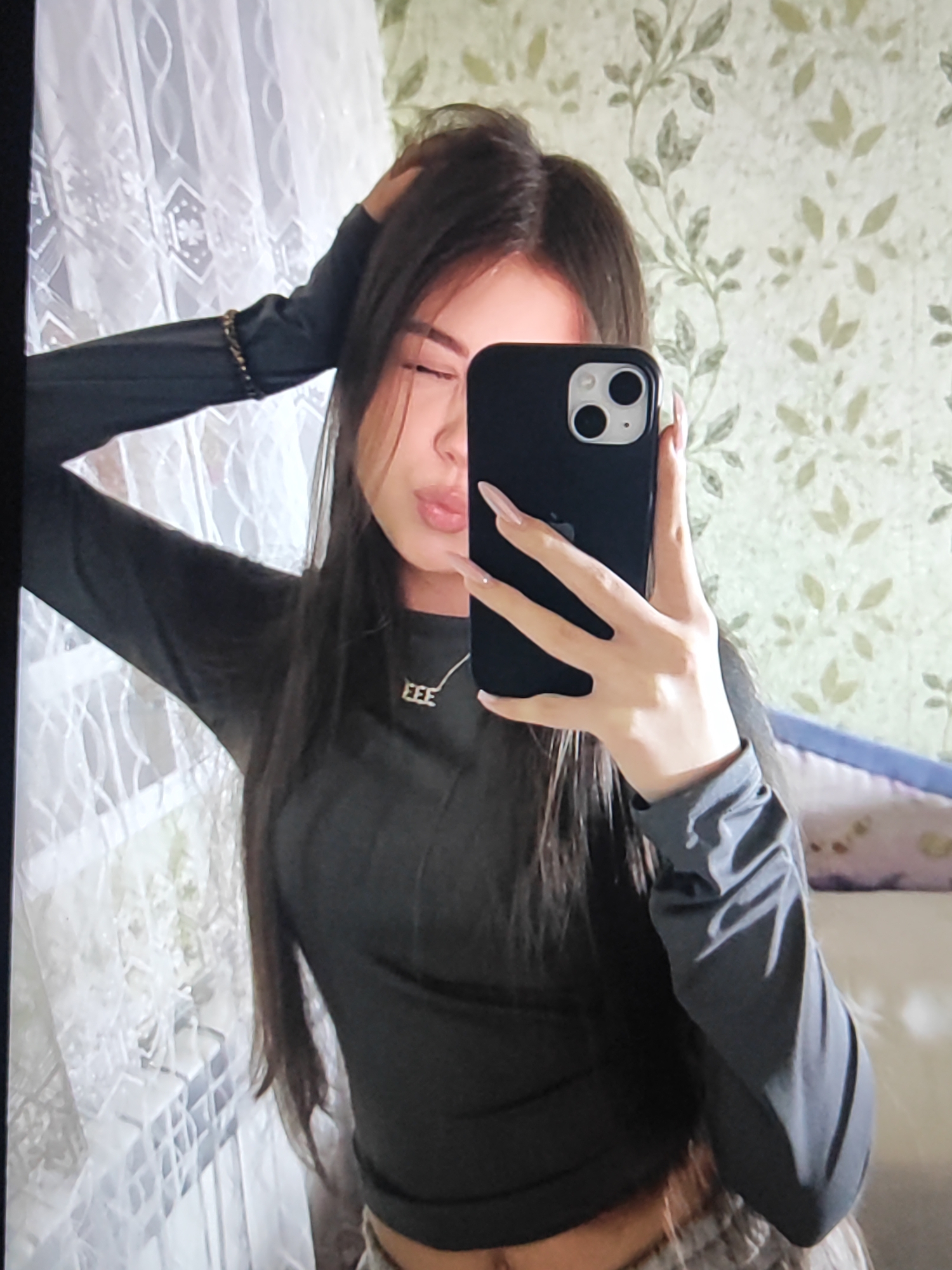 The image shows a young woman with long dark hair taking a mirror selfie. She is wearing a dark long-sleeved shirt and plaid pants. She has her left hand on her head and is holding her phone with her right hand to take the picture. She is wearing a necklace with a small pendant. The background appears to be a room with floral wallpaper and a window with a white curtain. The location is not discernible from the image provided.