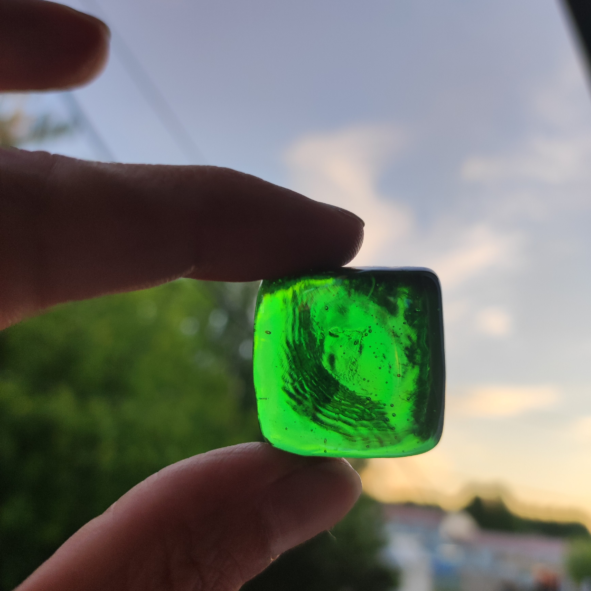 In Kineshma, Russia, at eye-level, a person's thumb and forefinger hold a small, translucent green, square-shaped glass object. The glass has visible swirls and small air bubbles within it. The background is softly blurred, showing a sky with scattered clouds, transitioning to a warmer tone near the horizon. Below the sky, out-of-focus green foliage is visible, followed by the indistinct shapes of buildings. The lighting suggests it is either sunrise or sunset.