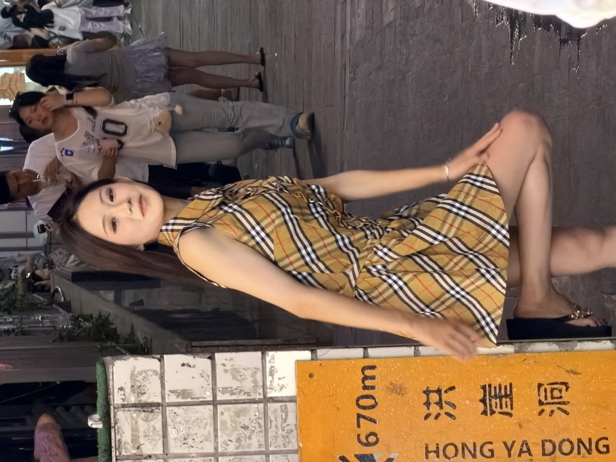 An East Asian woman is posed in front of a textured wall and a yellow sign on a city street. She is wearing a plaid dress and her left leg is bent at the knee. Behind her, other people are visible walking and standing. The yellow sign has Chinese characters and the text "670m HONG YA DONG". The street appears to be paved with wooden planks. The lighting suggests it is daytime.