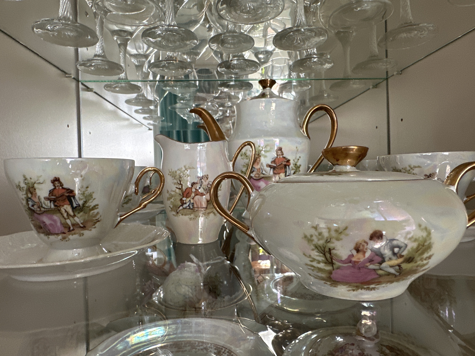 The image displays a collection of vintage porcelain teaware, including a teapot, creamer, and teacup with saucer, arranged on glass shelves within a display cabinet. The teaware is adorned with pastoral scenes featuring couples in period clothing. Above the teaware, several clear glass stemware items are inverted and hung from the top shelf. The cabinet's background is a light-colored wall, and the shelves are transparent glass, reflecting the items and the surroundings. The lighting appears to be ambient, highlighting the iridescent finish of the porcelain. No people are visible in the image. The location is unknown.