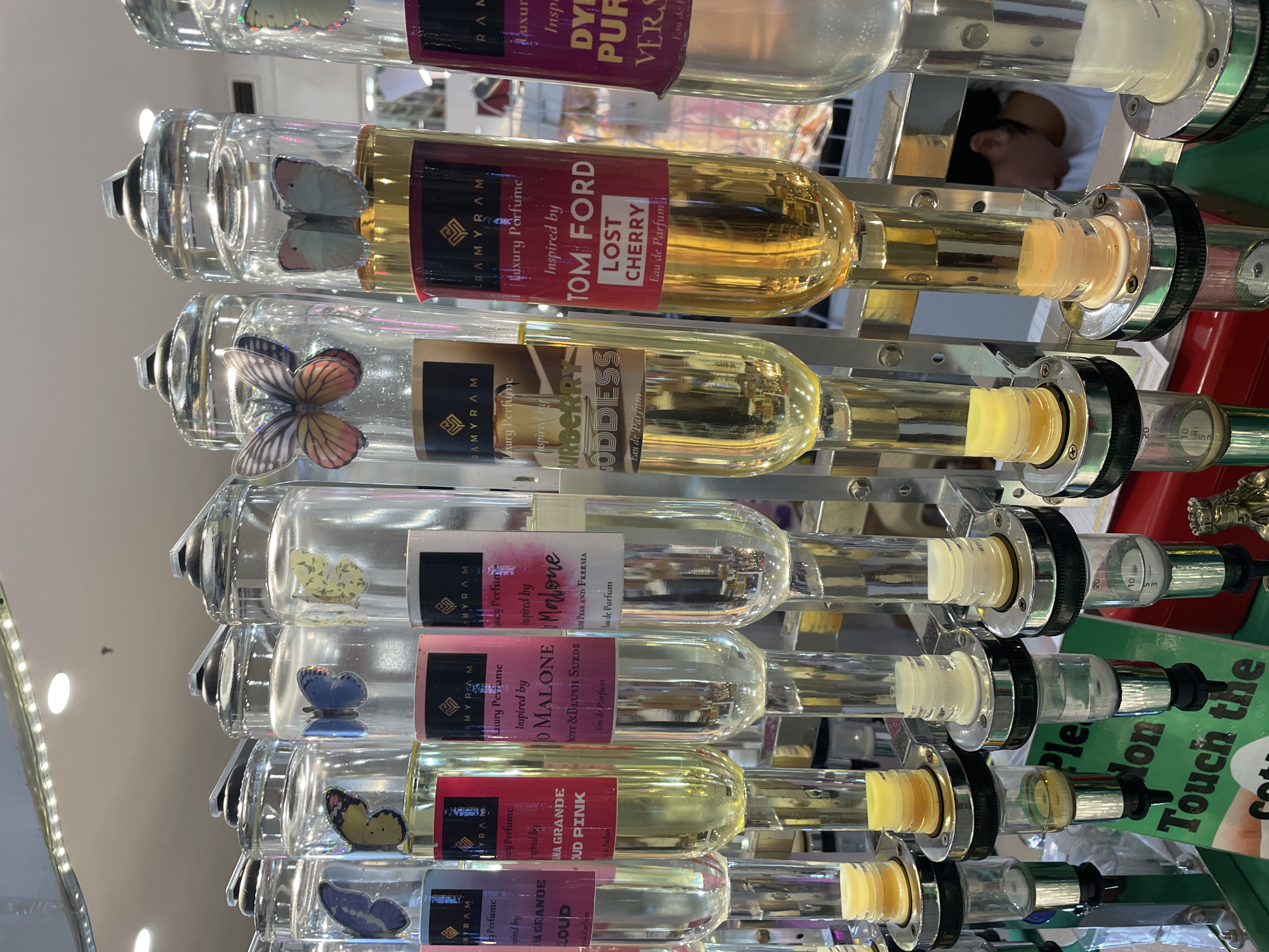The image shows a row of perfume dispensers with butterfly decorations, likely in a retail setting in Davao City, Philippines. The dispensers are made of glass and metal, each containing a different colored liquid and labeled with brand names and perfume inspirations, such as "MYRAM Luxury Perfume," "Inspired by Ariana Grande Cloud Pink," "Inspired by Jomalone Honey & Blushi Suede," "Inspired by Burberry Goddess," "Inspired by Tom Ford Lost Cherry," and "Inspired by Dyptique Pur." The dispensers have spigots at the bottom for dispensing the perfume. In the background, a person wearing a white shirt is visible, appearing to look down at the products. A green sign with white text partially visible reads "Please don't touch the..." and is positioned in front of the dispensers. The overall environment suggests a perfume shop or a market stall.