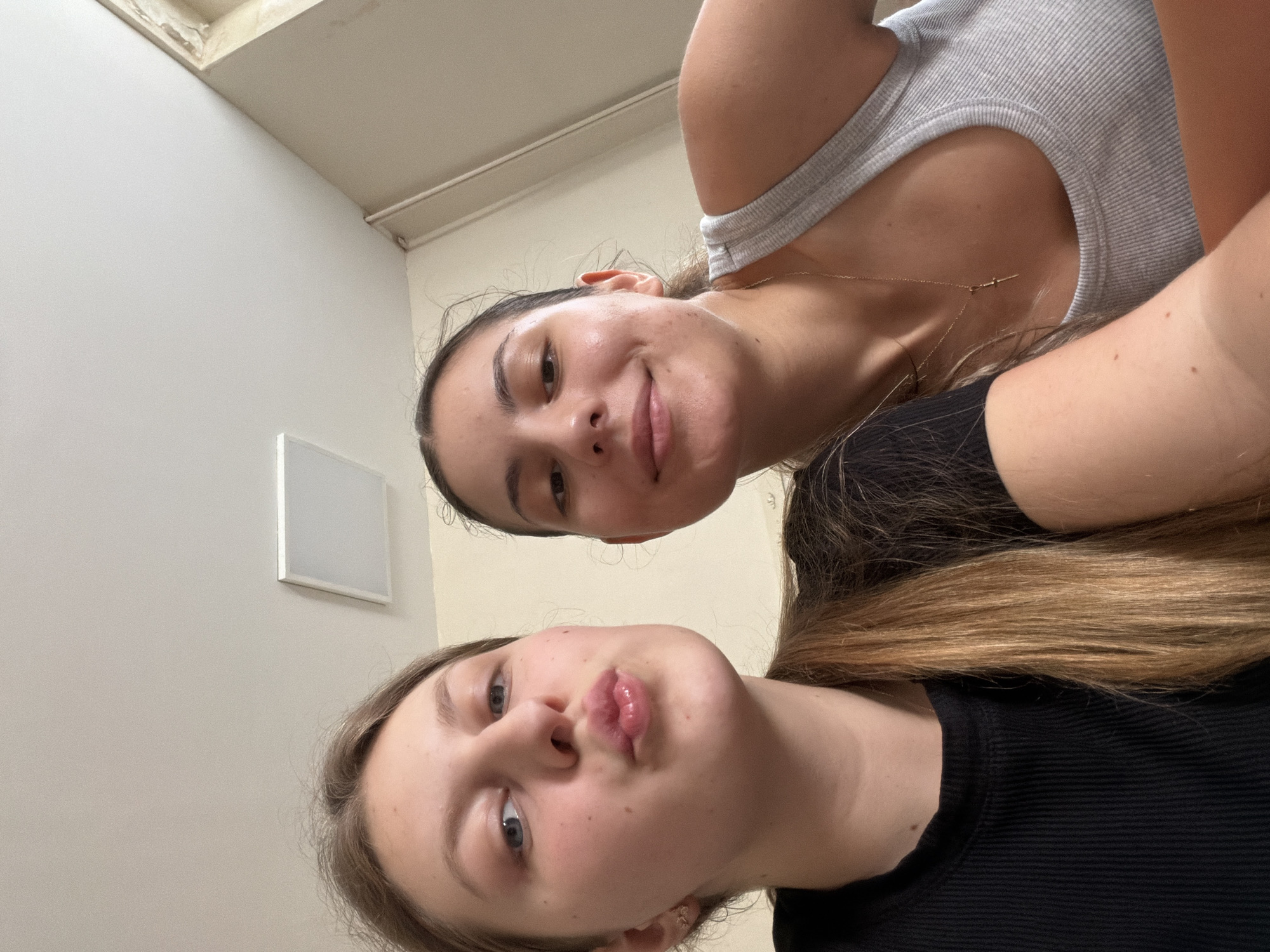 The image is a close-up selfie taken indoors, likely in an apartment or room, with two young women. The woman on the left has blonde hair and is making a kissing gesture with her lips. She is wearing a black ribbed t-shirt. The woman on the right has dark hair and is smiling. She is wearing a grey ribbed tank top and a delicate gold necklace with a cross pendant. The background appears to be a plain wall and ceiling with a square light fixture visible. There are no identifiable landmarks or elements that would suggest a specific location within Burgas, Bulgaria. The lighting is natural and comes from above.