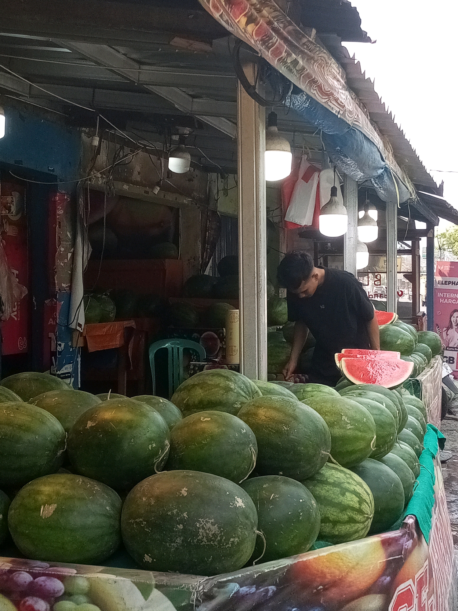 In Setiamekar, Indonesia, a young man in a black t-shirt is actively engaged in selling watermelons at an outdoor market stall. He is positioned behind a large display of watermelons, some of which are whole and others are sliced, revealing their red flesh. The stall is covered by a corrugated metal roof supported by visible beams, and adorned with patterned fabric. Several bright, dome-shaped lights are suspended from the ceiling, illuminating the scene. Behind the man, a variety of items are visible, including what appears to be shelving, stacked objects, and a green plastic chair. In the foreground, more watermelons are piled high, and a portion of a colorful banner depicting fruit is visible at the bottom edge of the frame. The overall environment suggests a bustling, open-air marketplace.