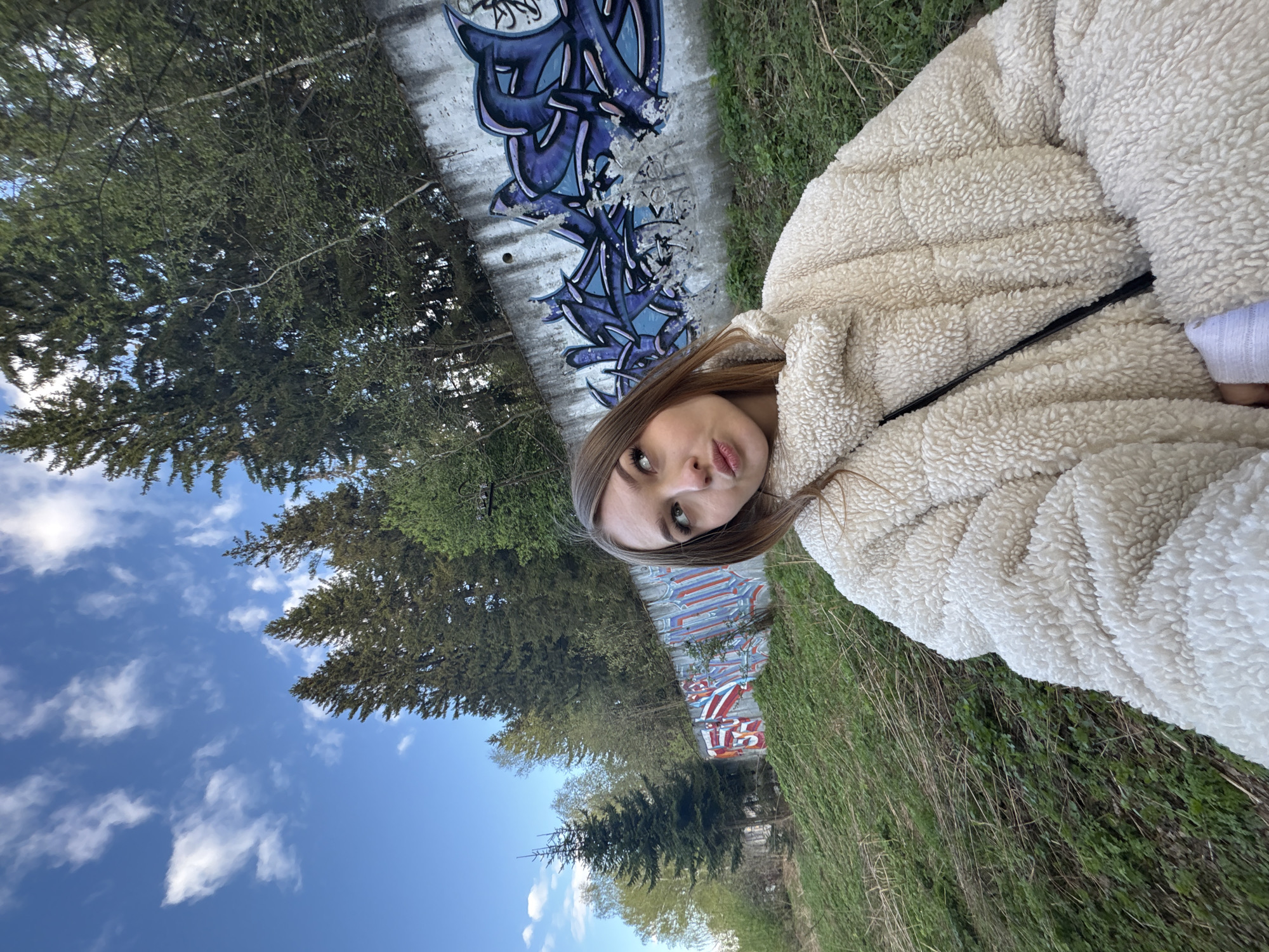 A woman with long brown hair is standing outside, near what appears to be a park or recreational area in Chaikovsky, Russia. She is wearing a cream-colored fleece jacket. A graffiti-covered wall and green vegetation run alongside her. In the background, there is a blue sky with white clouds and several trees. The image appears to be taken with a mobile phone.