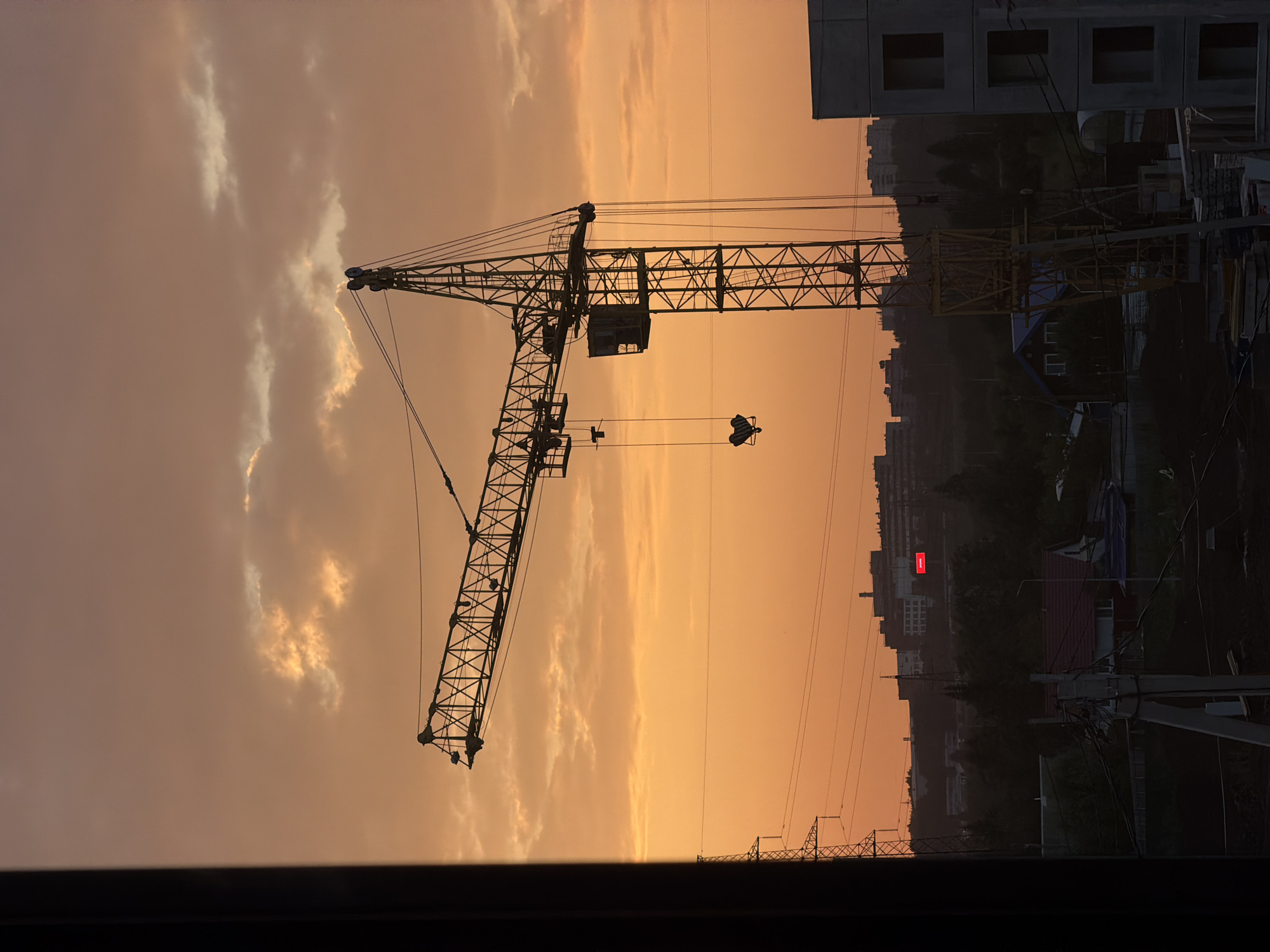 The image depicts a construction site at dusk, set against a vibrant orange sky with scattered clouds. A large tower crane, silhouetted against the sky, dominates the center of the frame. The crane's boom extends horizontally to the left, with a hook hanging from its end. Below the boom, a cabin is visible on the crane's structure. In the foreground, the silhouettes of buildings under construction, utility poles, and trees are visible. In the middle ground, a cluster of buildings, including some with red illuminated signs, are discernible. The overall scene suggests ongoing development in an urban or suburban area. The image does not contain any people or indicate any specific actions. Based on the provided context, the location is identified as the city of Froly, Russia.