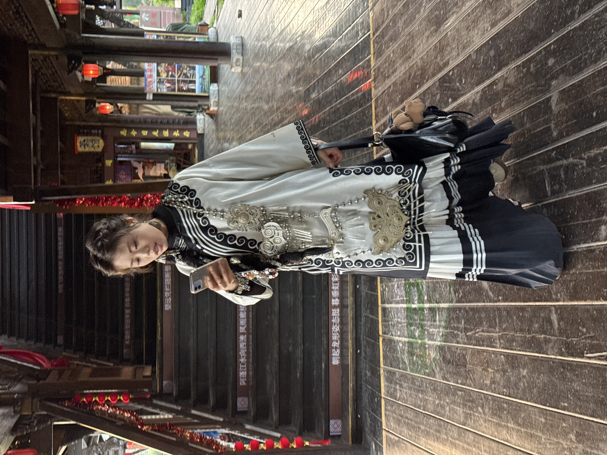 The image shows a woman standing in what appears to be an outdoor walkway or street, possibly in Zhuoshui, China, as indicated by the Chinese characters present on signage along the walkway. The woman is holding a smartphone and looking at it. She is dressed in what seems to be a traditional-style outfit with decorative silver accents and a black-and-white design. She also carries a small black purse or bag. The walkway is made of wooden planks, and there are wooden structures on either side, with hanging red lanterns. Steps lead down into an area with additional red decorations. The overall environment looks like a historic or tourist-oriented area.