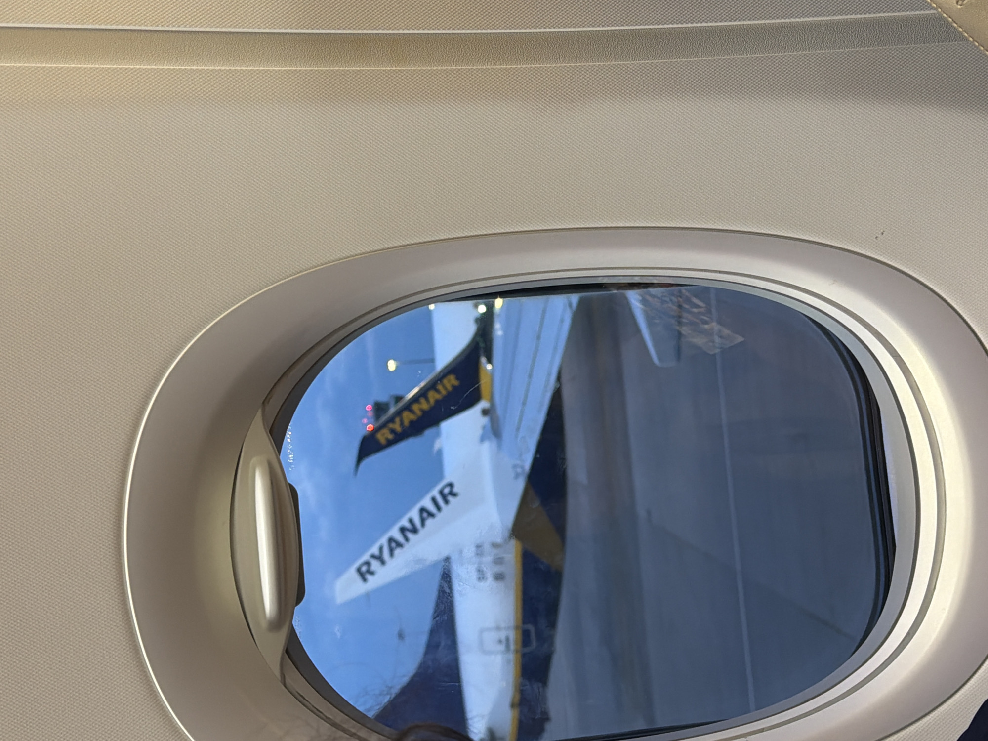 The image captures a view from inside an airplane, looking out through an oval-shaped passenger window. The immediate surroundings of the window, including the textured light beige interior paneling and the window frame, are prominently featured. A handle or latch mechanism is visible on the left side of the window frame.
Outside the window, a bright blue sky indicates daytime. The main subject seen is a reflection of another aircraft, likely a Ryanair plane, at Sofia Airport. The reflection clearly shows the tail fin of the other aircraft, which is dark blue with the word "RYANAIR" written vertically in white letters. Above the reflected tail fin, what appear to be red navigation lights are visible. Below this, a portion of the wing or horizontal stabilizer is reflected, also displaying the "RYANAIR" logo horizontally in white against a dark blue background. Below this horizontal text, some alphanumeric characters, possibly an aircraft registration starting with "SP-R", are partially visible. The reflections also suggest the presence of other airport structures or a jet bridge. The weather appears clear and sunny.