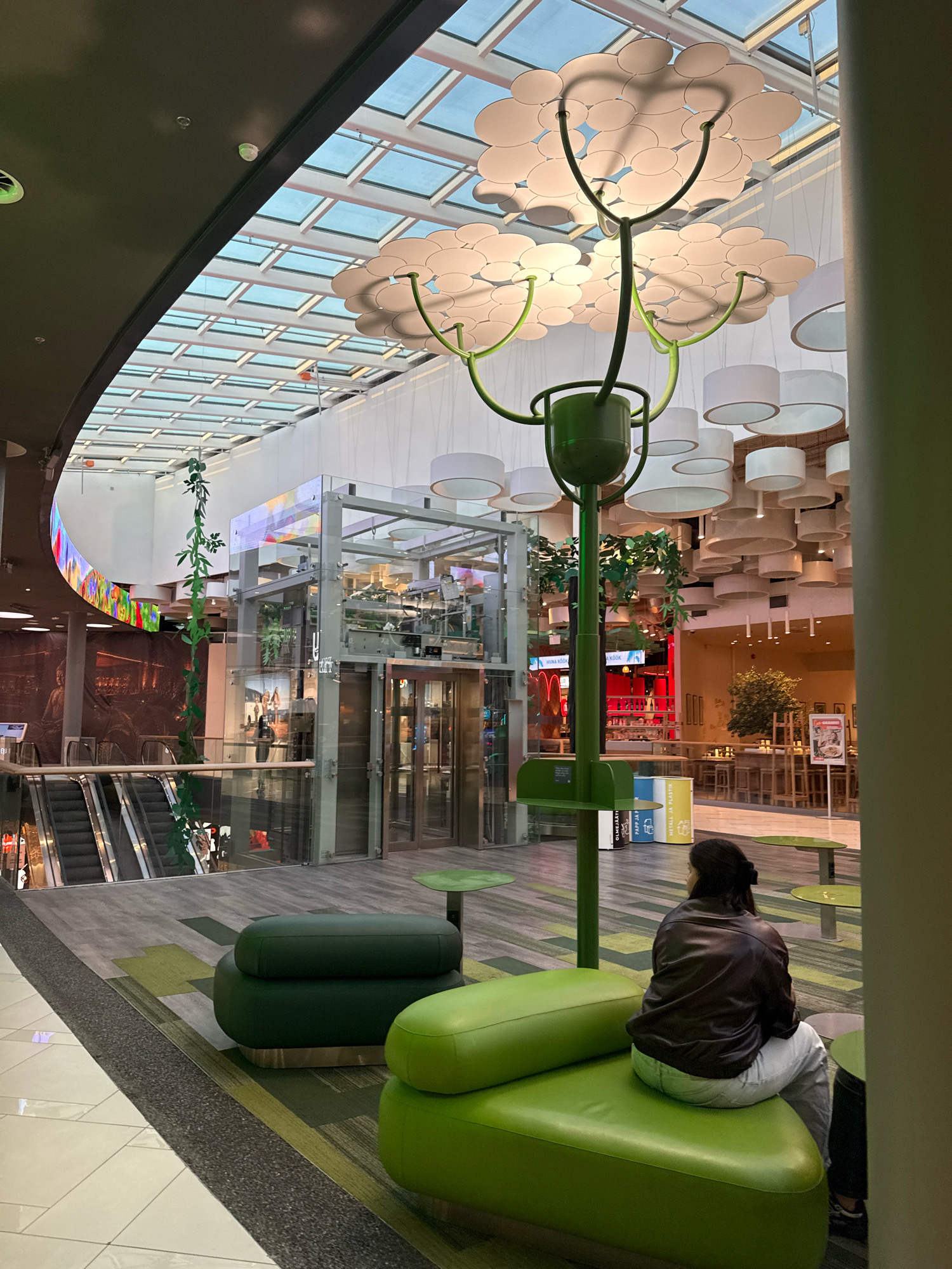 A young woman with dark hair pulled back is seated on a bright green, modular sofa in the atrium of a modern, indoor shopping mall. She is viewed from behind, wearing a dark brown leather jacket and light-colored pants, and appears to be looking into the distance. The sofa has a low profile and is upholstered in a shiny, smooth material. The floor in front of the sofa is a patterned carpet in shades of green and gray, with a dark gray border. To the left of the sofa is a dark green ottoman. In the background, a glass elevator enclosure is visible, along with escalators. The ceiling of the mall features a large, decorative light fixture resembling a tree with circular white lamps, and a glass roof allows natural light to enter. The environment suggests a public space, likely a commercial center, with elements of contemporary design and artificial greenery. This image was captured in Tallinn, Estonia.
