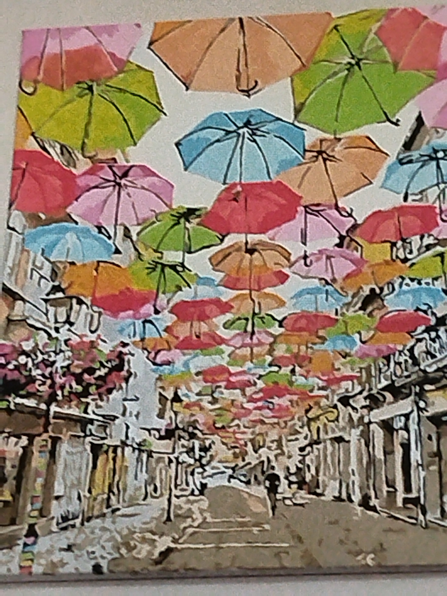 The image depicts a street scene, likely an art installation or festival decoration, where numerous colorful umbrellas are suspended overhead, creating a vibrant canopy. The umbrellas, in shades of pink, green, orange, and blue, are arranged densely across the street, partially obscuring the view of the sky. Below the umbrellas, the scene shows buildings with shops or businesses lining both sides of a street. The architecture appears somewhat traditional, with windows and storefronts visible. A single figure, silhouetted and small in the frame, is walking down the street, suggesting a sense of scale and human presence within the colorful display. The ground is paved, and there are indications of pavement and possibly cobblestones. The overall impression is one of a lively and decorative urban environment. There is no specific indication or context within the image that places it in Verkhnodniprovsk, Ukraine.