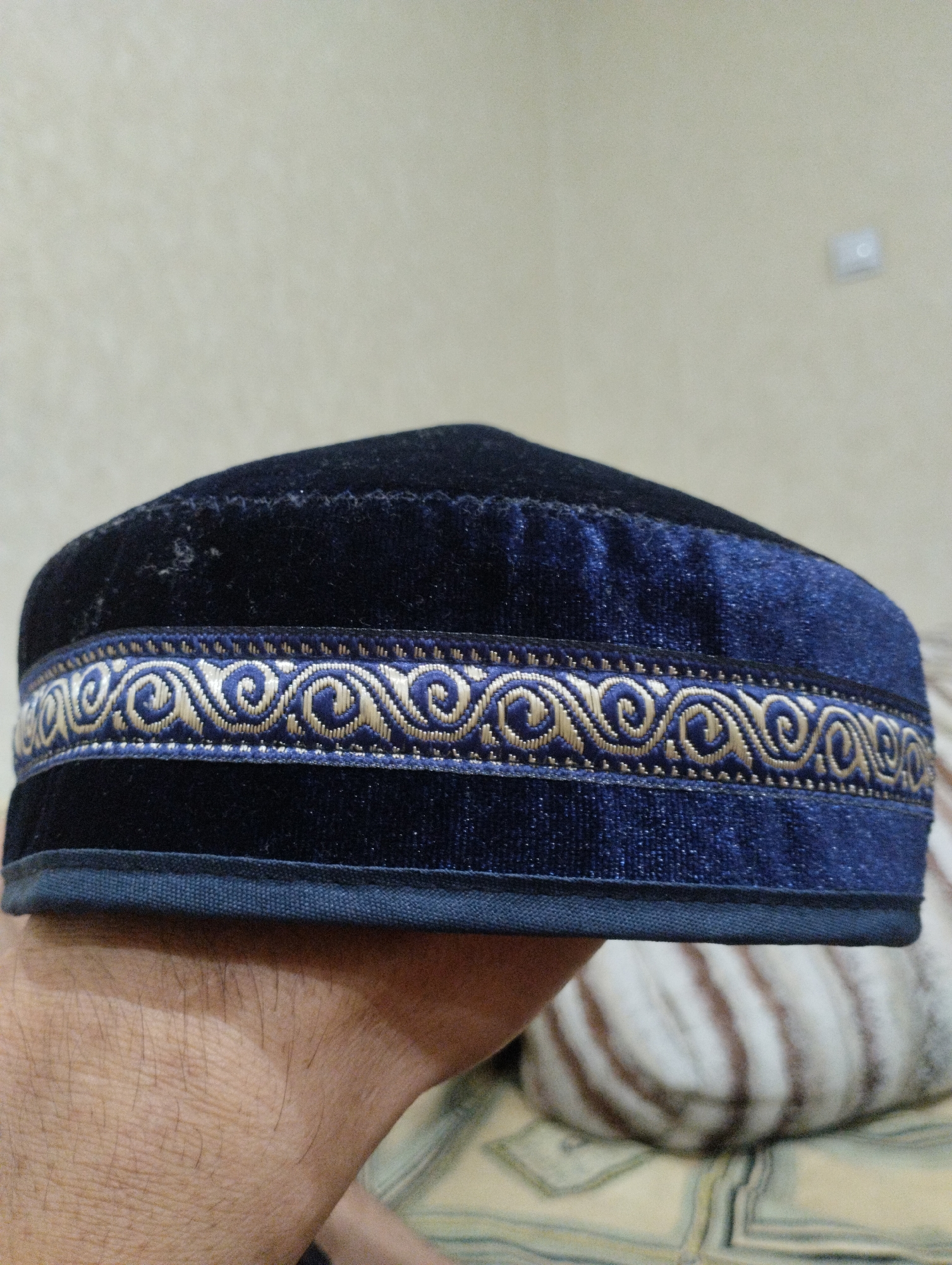 A dark blue velvet kufi cap with a golden and blue patterned trim is held on a person's hand. The cap is being presented or displayed from a close-up, low-angle perspective. The background consists of a plain, light-colored wall and a patterned surface, possibly a blanket or rug, with soft, out-of-focus elements. The scene appears to be indoors, in a residential setting. There are no identifiable landmarks or features that would suggest a specific location in Semey, Kazakhstan.