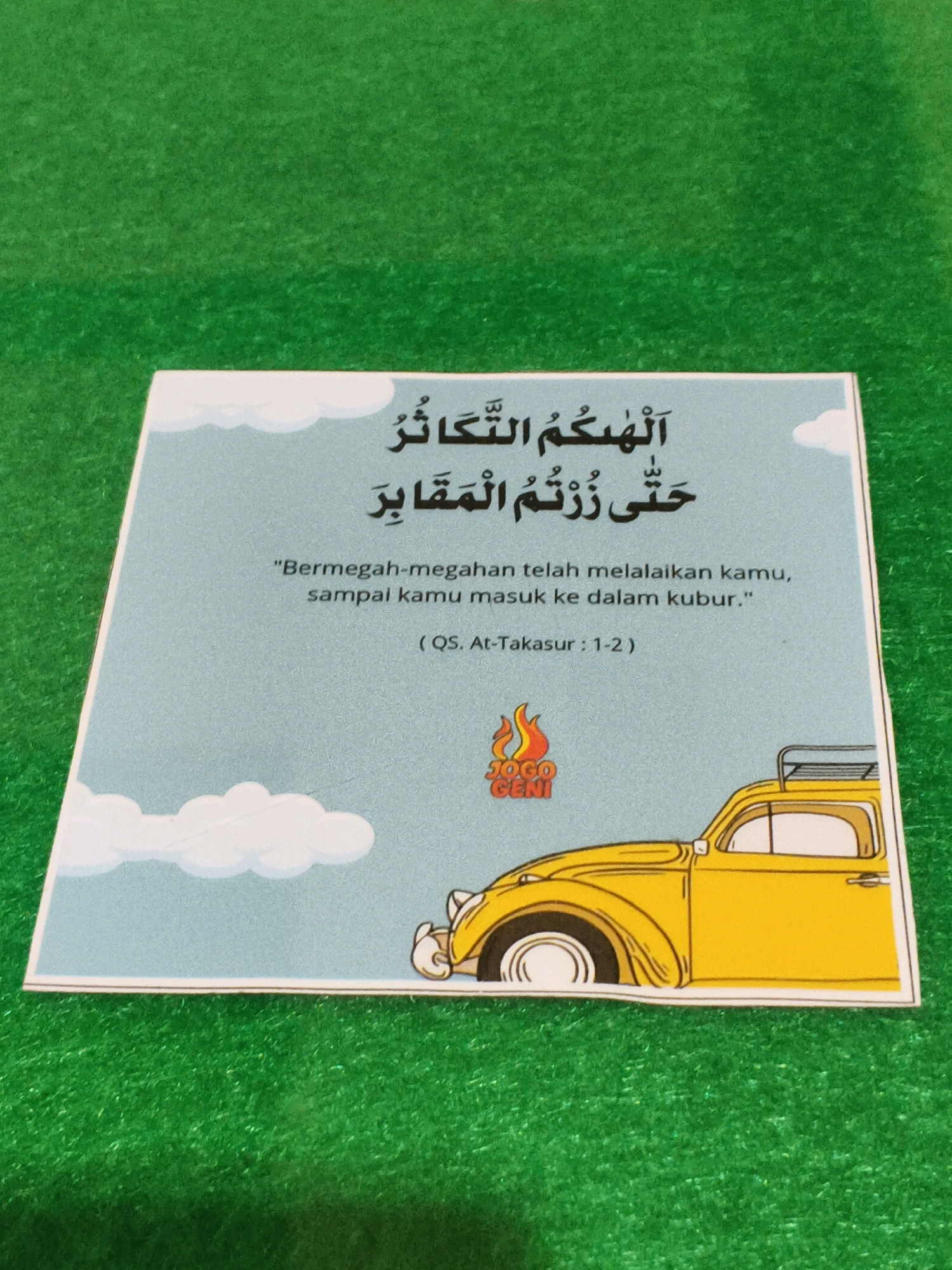 A religious postcard with Arabic text and an illustration is placed on a green, textured surface, resembling artificial grass. The postcard has a light blue background with white cloud graphics. The Arabic text, written in black, is positioned at the top of the postcard. Below the Arabic text, there is a horizontal line followed by Indonesian text in quotation marks, translating to "Being rich-rich has distracted you, until you enter the grave." Beneath this, the reference "(QS. At-Takasur : 1-2)" is provided.
In the lower right corner of the postcard, there is a cartoon illustration of a yellow vintage car with a roof rack. To the left of the car, there is a logo that reads "JOGO GENI" with a flame design.
The green surface the postcard rests on appears to be consistent with the carpeting found in a home or a community space, such as a children's play area or a religious study room, common in residential neighborhoods like those found in Karangmalang, Indonesia. The lighting suggests an indoor environment with ambient light. There are no people or animals visible in the image. The focus is solely on the postcard and its immediate surroundings.