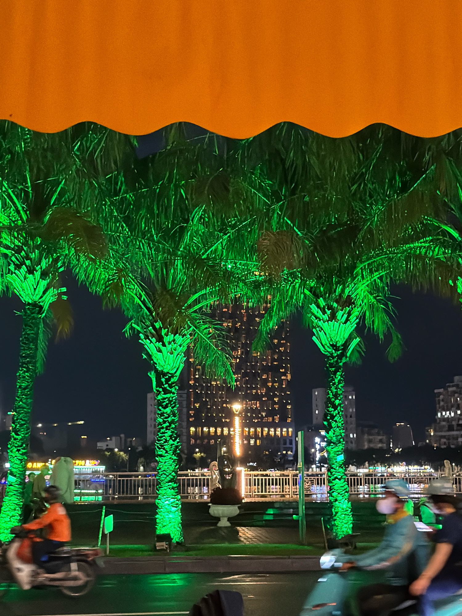 The image captures a nighttime scene in Da Nang, Vietnam, likely along the Han River. Several palm trees are illuminated with vibrant green lights, creating a striking contrast against the dark sky. In the background, a tall, modern building with numerous illuminated windows dominates the skyline, suggesting a bustling urban environment. A bridge or embankment with railings and decorative lights is visible, spanning across the river. On the road in the foreground, two individuals are riding on a scooter. The person in front is wearing an orange top and a helmet, while the person behind is wearing a blue jacket and a mask. The road surface appears wet, reflecting the green illumination from the palm trees. The overall atmosphere is festive and energetic, indicative of a city that embraces its nighttime attractions.