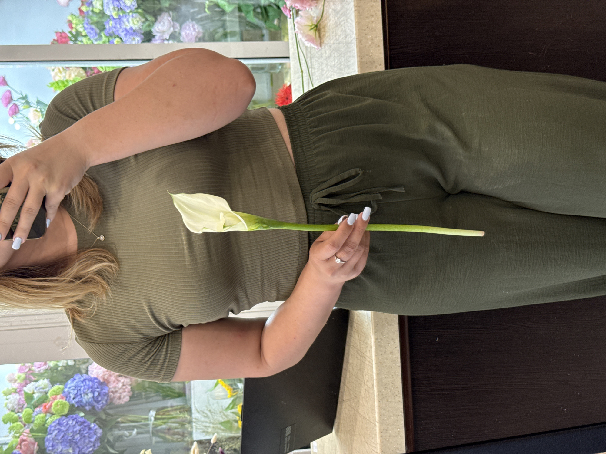 A woman is standing in what appears to be a room with large windows, possibly a flower shop, given the abundance of flowers visible in the background. She is wearing a green, ribbed crop top and matching green, wide-leg pants. Her hair is blonde and falls to her shoulders. She is holding a single white calla lily in her right hand. Her left hand is holding a smartphone, implying she might be taking a selfie or recording a video. A laptop is visible on a counter to her left. The overall lighting suggests an indoor setting during the day.