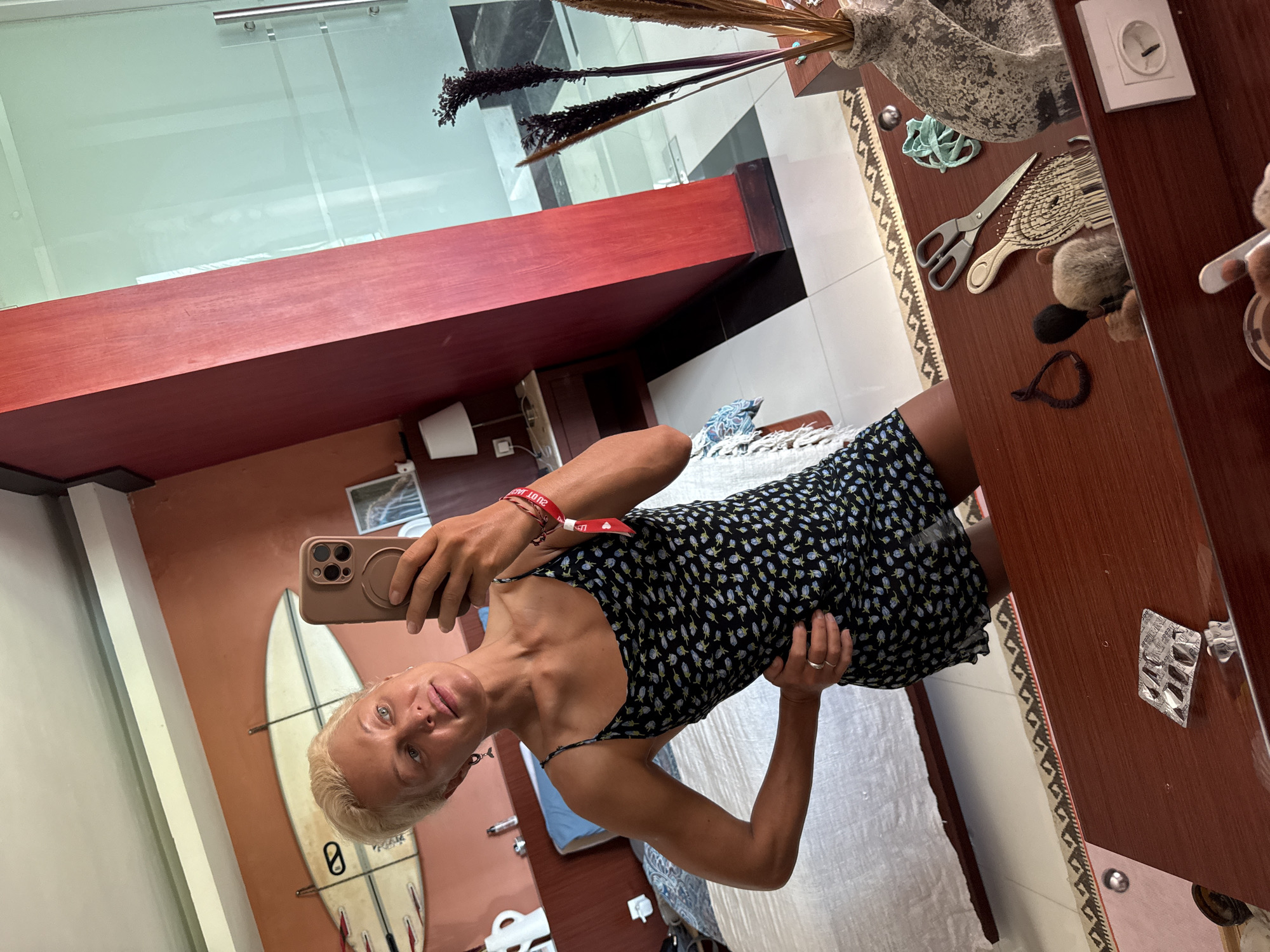 In a room with coral-colored walls, a woman with short blonde hair stands in front of a mirror, taking a selfie. She is wearing a black slip dress with a floral pattern and has a red wristband on her left arm. Her left hand is on her hip, and her right hand holds a smartphone. Behind her, a bed is visible, with surfboards mounted on the wall above it. To her right, a large red pillar stands next to a glass-enclosed shower. The mirror's surface reflects the room, including a dresser with various items such as scissors, a vase with dried flowers, and a blister pack of pills. The floor is tiled with black and white squares.