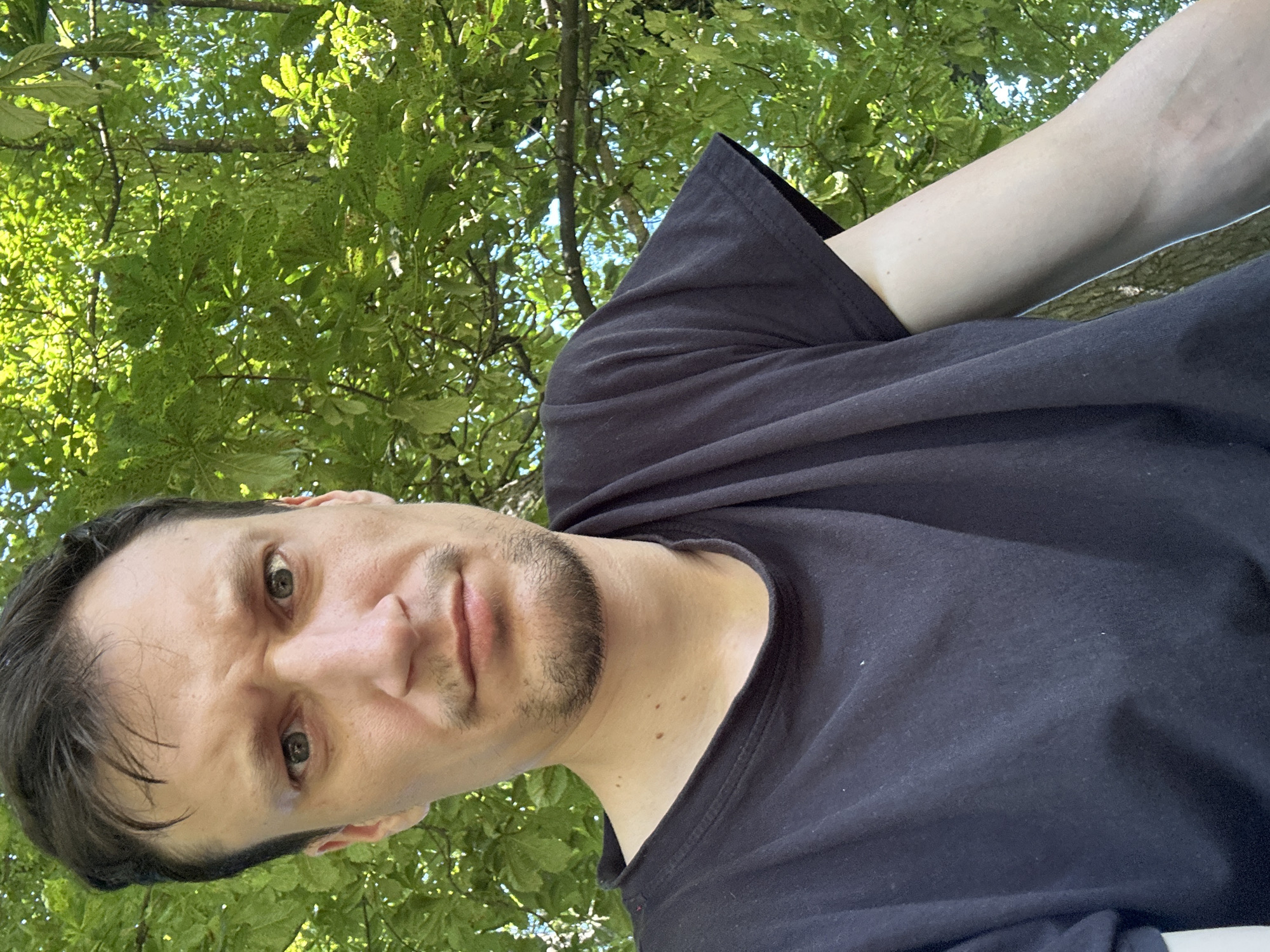 The image is a close-up selfie of a man with dark hair and a short beard. He is wearing a black t-shirt and is outdoors, with green trees and foliage in the background. The man's expression is serious, with his eyebrows furrowed. His left arm is visible, resting on his chest. The lighting suggests it is daytime, possibly sunny, casting some shadows. The image does not provide any specific details that would allow for its location to be determined within the city of Makiv, Ukraine.