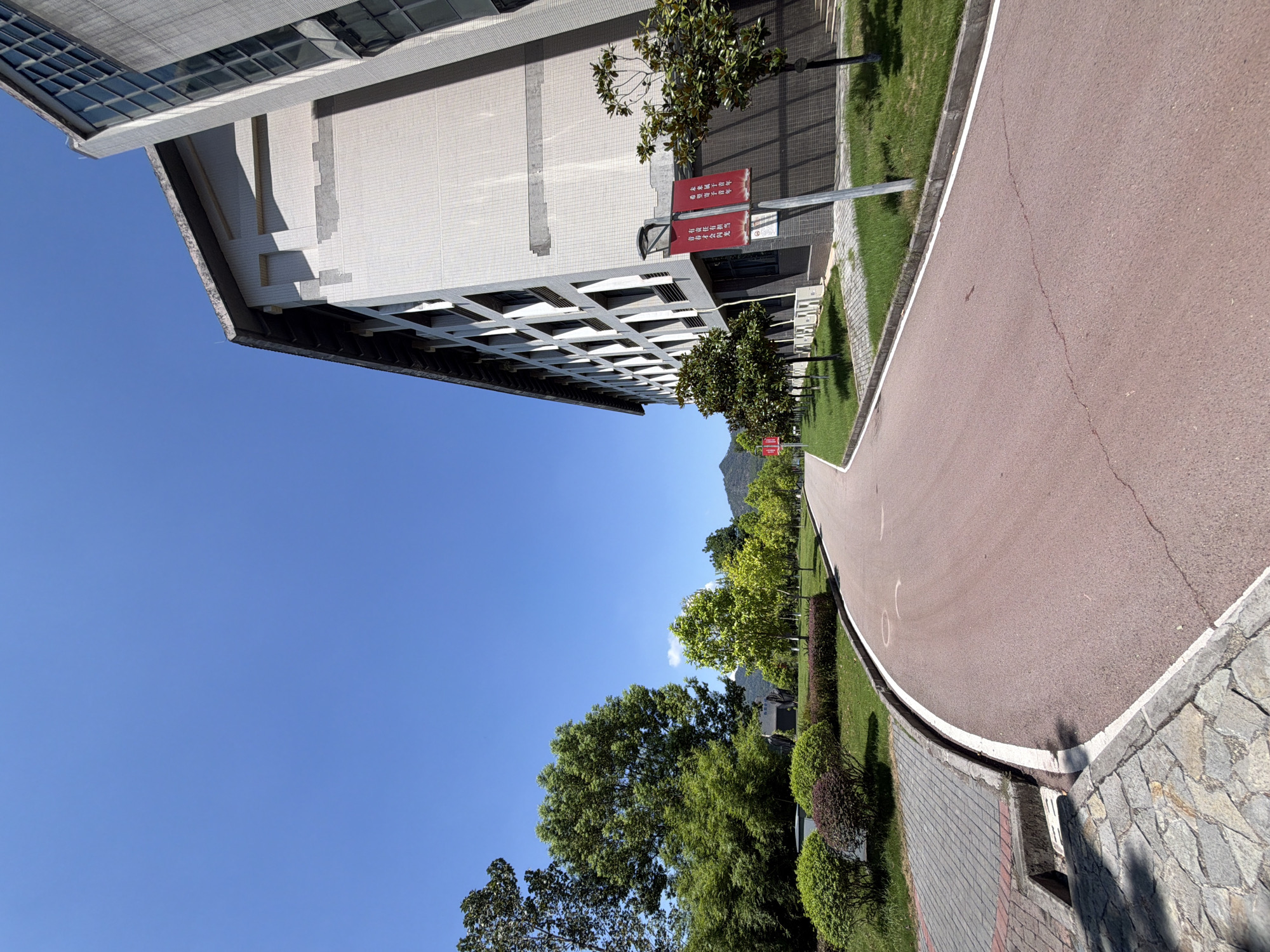 This image depicts an exterior view of a modern building with a light-colored tiled facade, located in Jiangjin, China. The building is flanked by a paved pathway and a well-maintained grassy area with trees and shrubs. A reddish-brown asphalt road curves into the distance, bordered by a white line. The sky is clear and blue, with a hint of sunlight casting shadows. Two red banners are affixed to a pole near the building, featuring Chinese characters. The architecture suggests a public institution or a commercial complex, typical of urban developments in China.