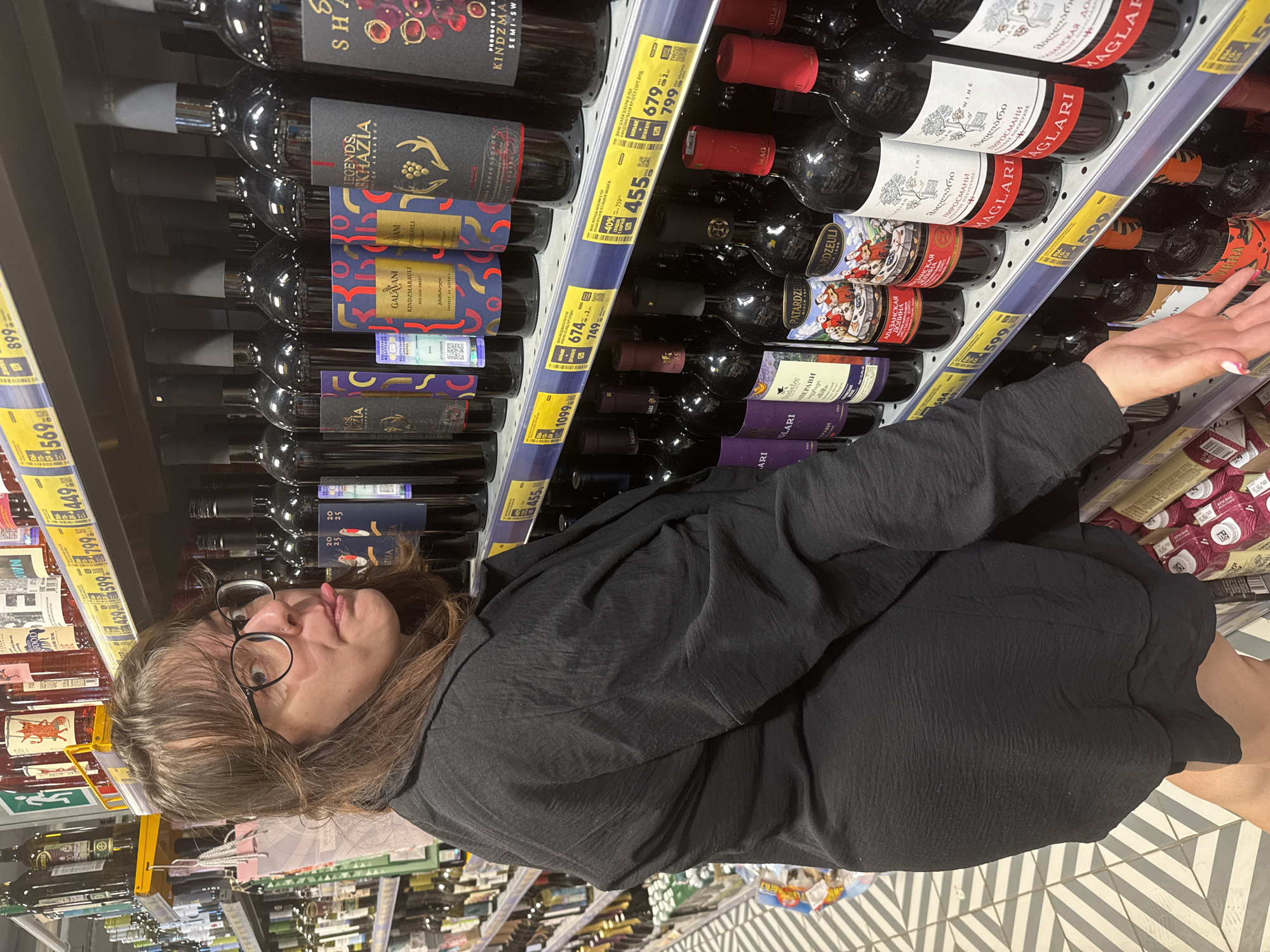 In Dmitrov, Russia, a woman with brown hair, glasses, and a black textured jacket stands in a supermarket aisle. She is sticking her tongue out at the camera. The aisle is lined with shelves stocked with bottles of wine. The shelves are equipped with yellow price tags. The floor is tiled with a black and white geometric pattern.