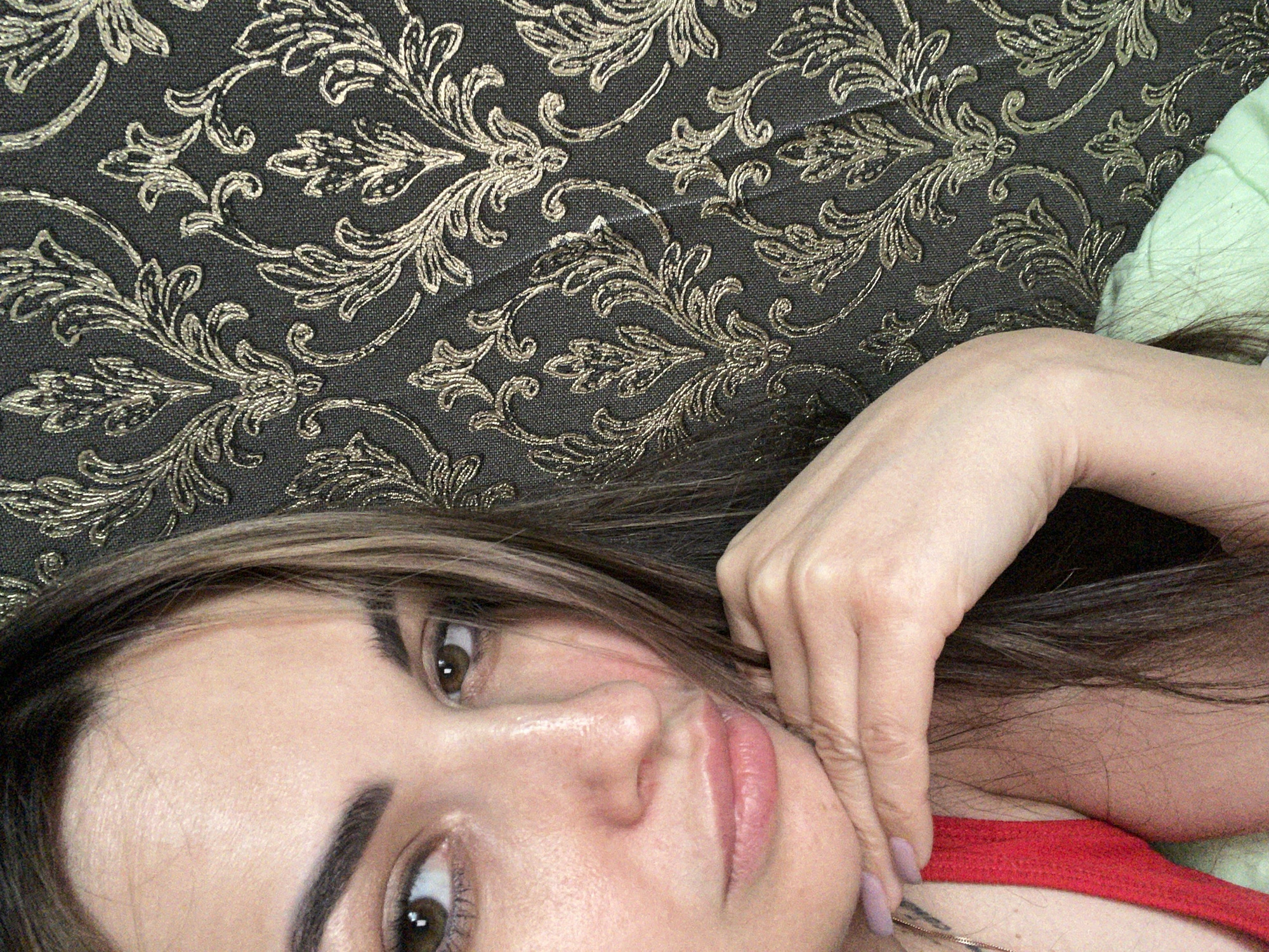 A close-up, half-body portrait features an adult female positioned off-center to the left of the frame. Her head is tilted slightly, and her left hand (viewer's right) is supporting her chin, with fingers curled and resting against her jawline. Her fingernails are painted a light lavender hue. She has long, multi-tonal brown hair with lighter highlights, which partially covers the right side of her face and falls over her shoulders. Her brown eyes are visible, with her gaze directed towards the left side of the image. Her eyebrows are dark and well-defined. Her lips are full and appear a natural light pink. She is wearing a red garment, possibly a strap, visible on her left shoulder, and a light green garment is visible on the lower right portion of the image. A delicate gold-colored chain necklace is visible on her neck. The background consists of a dark, possibly charcoal grey or black, surface with a raised, intricate metallic gold or bronze damask pattern featuring elaborate scrollwork and floral motifs. The texture of the background suggests wallpaper or a similar decorative textile. The scene is located in Unknown, Unknown.