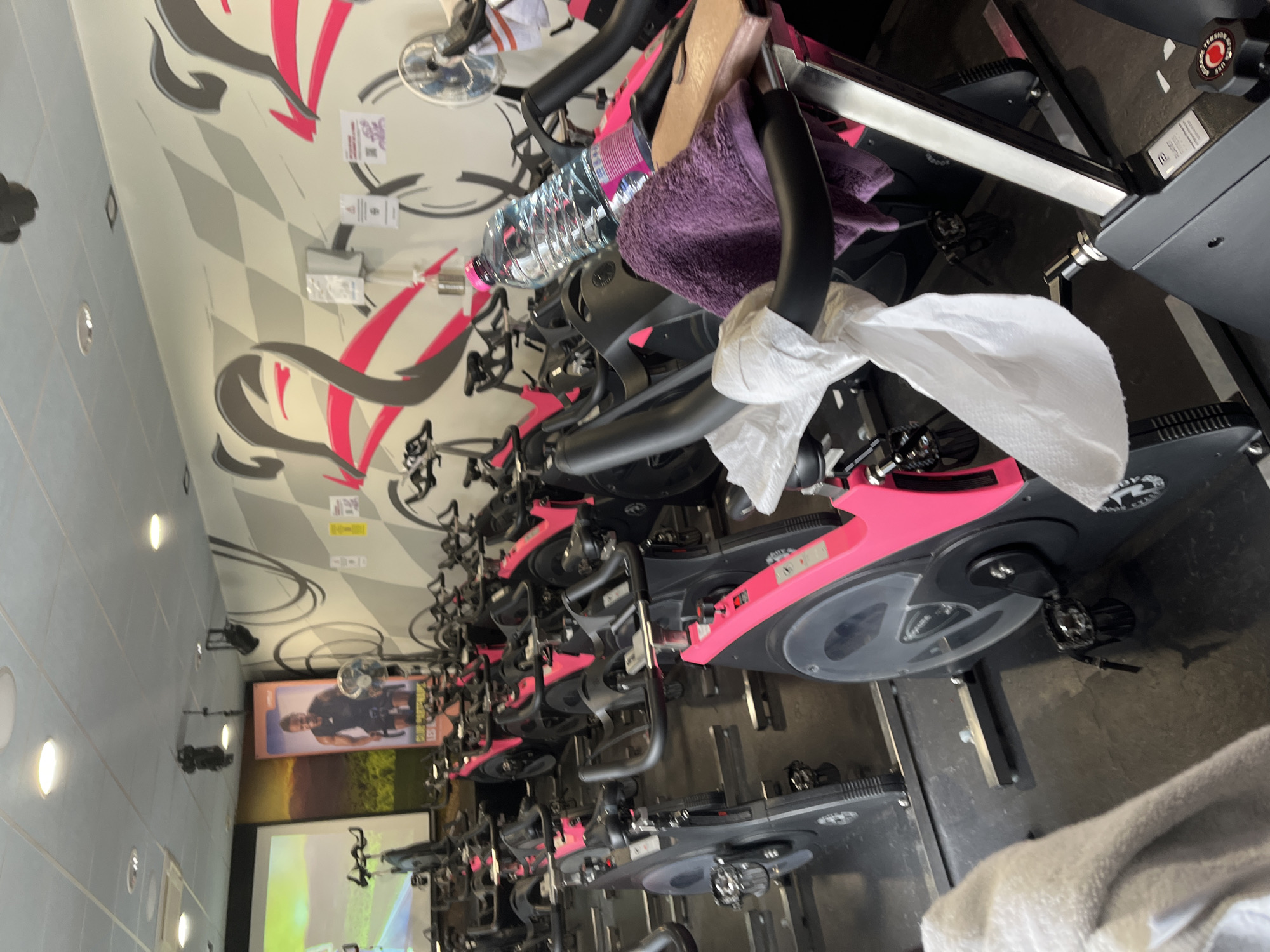 The image shows a fitness studio in Saint-Galmier, France, filled with stationary bicycles. The bikes are mostly black and pink and arranged in rows. Some bikes have towels and water bottles on them. The studio has a modern design with gray and pink accents on the walls. A screen displays a cycling video. There are no people visible in the immediate foreground.