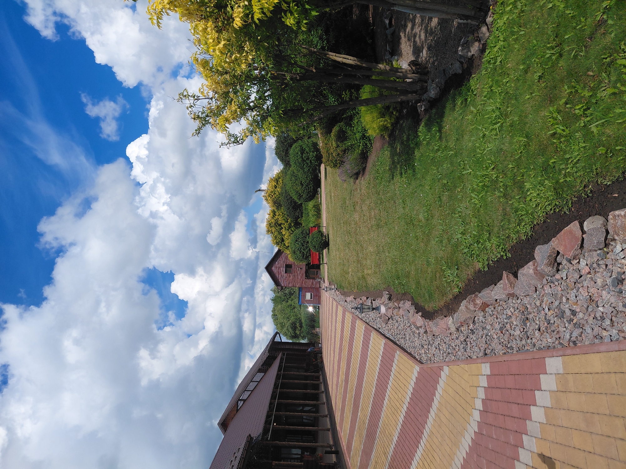 The image depicts a view of the resort complex "Knyazhyi Dvir" in Kovalivka, Ukraine, on a sunny day with scattered clouds. In the foreground on the left, a paved walkway made of red and yellow rectangular tiles runs diagonally towards the background. The walkway is bordered by a strip of grey gravel and large reddish-brown stones. Beyond the gravel border, a well-maintained green lawn stretches into the middle ground, with a small garden bed featuring lavender and other low-lying plants.
To the left of the lawn is a long, wooden building with a dark brown roof, featuring a covered porch supported by thick wooden columns. A person is visible near the building in the middle ground, seemingly tending to something on the ground.
In the middle ground, to the right of the lawn, stands a small, dark red wooden building with a darker roof. Adjacent to this building are two bright red benches. Further back, a variety of green bushes and trees are visible, including a tree with bright yellow foliage in the upper right portion of the image. The sky above is a vibrant blue with numerous white, fluffy cumulus clouds. The overall scene conveys a sense of a well-kept outdoor recreational or hospitality area.