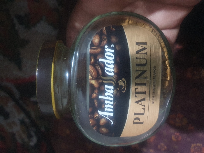 The image displays a glass jar of instant coffee, held horizontally by a person's hand. The jar is filled with light brown, granulated coffee, visible through the clear glass. A prominent label wraps around the jar, featuring a dark background adorned with an image of roasted coffee beans.
Visible text on the label includes the brand name "Ambassador" written in a white, stylized script font on the left side, accompanied by a registered trademark symbol (®) and a small coffee cup icon. To the right of the "Ambassador" logo, the word "PLATINUM" is vertically displayed in large, gold, sans-serif capital letters. Below "PLATINUM," the phrase "RICH SMOOTH FLAVOR" is written in smaller text.
The jar has a dark brown or black lid, with a thin yellow or gold band visible just beneath it. The background of the image is dark and out of focus, showing indistinct patterns that suggest an indoor setting, possibly on a patterned fabric or carpet. Part of a hand, with fingers visible, is holding the jar. No specific time of day or other environmental details are discernible from the image itself. The photograph provides a clear view of the coffee product.
