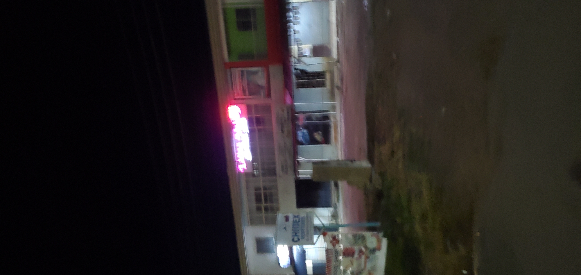 This blurry, low-light photograph depicts a night-time street scene in Gura Top, Nigeria, featuring commercial buildings. The sky is dark, indicating late evening or night.
In the midground, a two-story building complex is visible. The left section of the complex is a light-colored (likely white or grey) building. On its ground floor, a prominent white sign with blue text, "CHIDEX VENTURES", is mounted on a light blue pole structure. Below "VENTURES", additional smaller, illegible text is present. To the immediate left of this sign, a smaller, obscured white or light blue illuminated sign is partially visible. The upper floor of this section features multiple windows.
The central and right sections of the building complex include a vibrant pink and purple neon sign on the upper floor, clearly displaying the word "TUMBLAND". Below this sign, more windows are present. To the far right, the building's facade changes, featuring a distinct red section below a green section with a window. The ground floor of the central and right buildings appears to house several shopfronts, some dimly lit or dark, with reflections or faint interior details visible in some windows.
The foreground consists of a dark ground surface, likely a road or unpaved shoulder, with indistinct, dark vegetation visible on the left side. A light-colored, rectangular concrete block or pillar stands vertically in the center-midground, slightly to the right of the "CHIDEX VENTURES" sign. Faint horizontal lines, possibly power lines, are visible in the dark sky above the buildings. No individuals or vehicles are visible in the scene. The overall image is soft and lacks sharp focus due to the low light conditions.