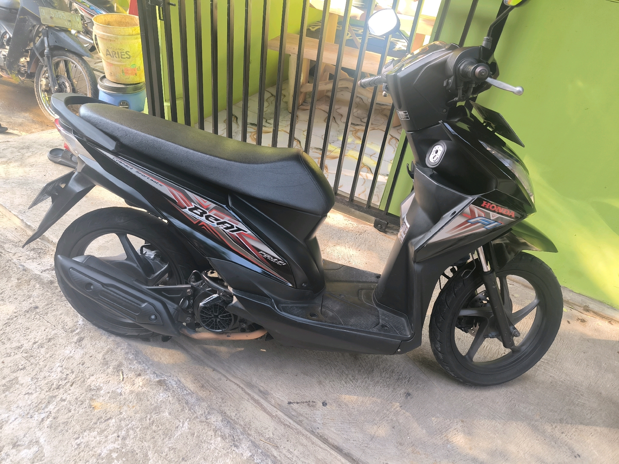 A black Honda Beat scooter is parked on a concrete ground next to a green wall with vertical black metal bars. A yellow bucket with red text that reads "ARIES" is visible behind the scooter, and to the left, another motorcycle is partially visible. The scooter's seat is black, and it has red and silver decals on its side panels. The front wheel and part of the engine are also visible. The sun is casting shadows on the ground, indicating it is daytime. The scooter appears to be in good condition. The scene is likely in a residential area or a small business in Cilengkrang, Indonesia.