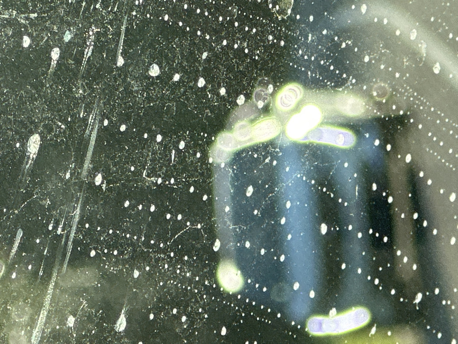 The image depicts an obscured view through a transparent surface, likely a vehicle windshield or window, heavily coated with numerous small, white, irregularly shaped particles or droplets, consistent with water and dust, along with fine linear scratches and grime patterns. Behind this foreground obstruction, a severely blurred and out-of-focus scene is visible. A dark-colored vehicle, potentially blue or black, is vaguely discernible in the center-left. Multiple bright, ovoid, and circular light sources or reflections, exhibiting green, white, and purple hues with a strong bokeh effect, are scattered across the blurred background, particularly on the left and right. No individuals, specific actions, or direct interactions are visible. The environment is indistinct due to the heavy blur and foreground obstruction, but the presence of a vehicle and light sources suggests an outdoor setting in the city of Saulx-les-Chartreux, France.