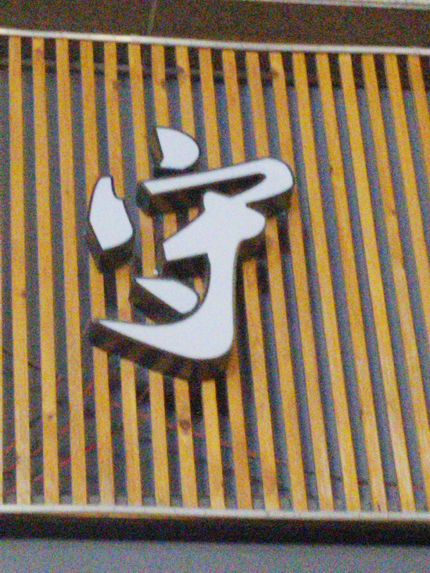 The image shows a close-up of a sign with a large, white, stylized Chinese character. The sign is set against a background of alternating vertical stripes, likely wood or bamboo, in shades of yellow and gray. The character is slightly raised, casting a small shadow on the striped background. The scene appears to be a detail of signage, potentially from a shop or restaurant. The Jiulongpo District location is inferred, not directly visible.
