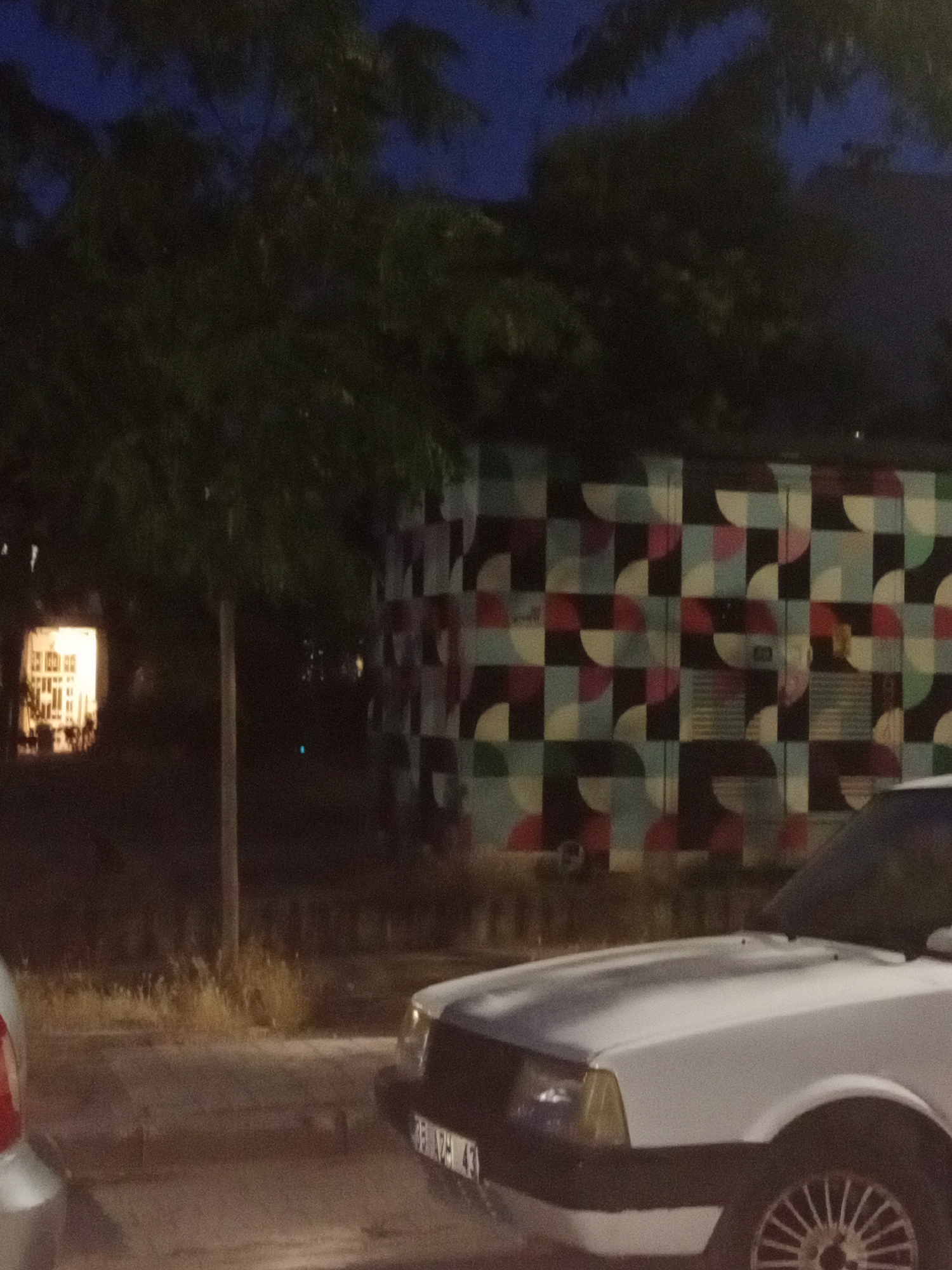 The image depicts a nighttime scene in Karşıyaka, Turkey. A white car is prominently featured in the foreground, partially obscuring a sidewalk and a street. Behind the car, a building with a distinctive geometric mural is visible. The mural is composed of squares and semicircles in various colors, including black, white, light blue, and shades of red and pink. To the left of the building, a brightly lit window suggests an indoor space.
The setting appears to be urban, with trees lining the area and dry vegetation along the roadside. The sky above is dark, indicating nighttime. The overall mood of the image is quiet and urban.
There are no clearly discernible people in the image, although faint shapes that could be figures are present within the building behind the mural. The activity taking place is not evident, as the scene is static.
Visible text includes a license plate on the white car, but the characters are too blurry to be read. Another faint text appears on the mural itself, but it is illegible. The time of day is clearly night, as evidenced by the dark sky and artificial lighting. There are no direct cues about the weather, but the general atmosphere suggests a clear night.