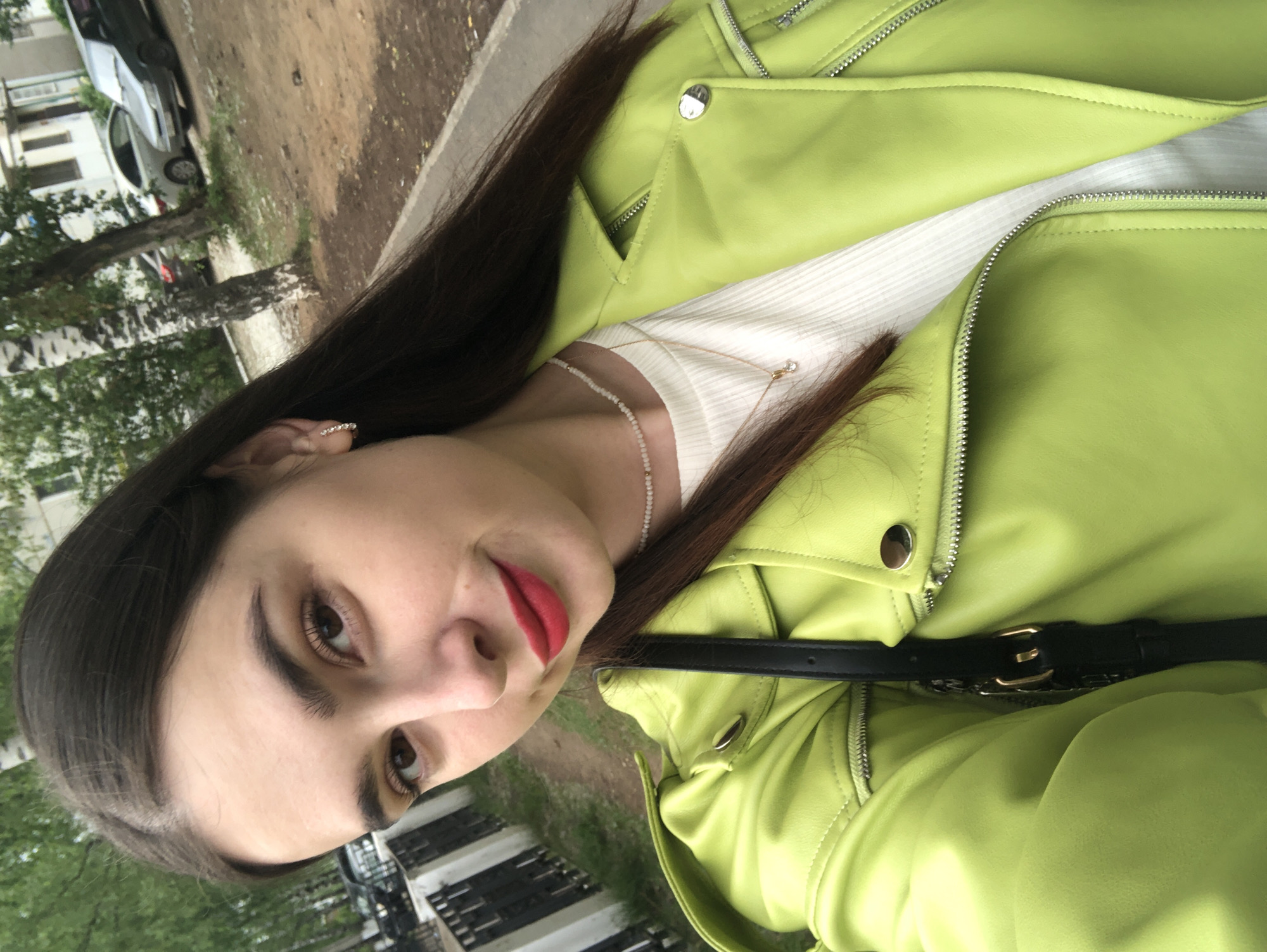 This is a waist-up, slightly angled selfie of a young woman with dark brown hair, taken outdoors. She is wearing a bright lime green faux leather jacket over a white ribbed top. A thin, delicate necklace with small beads and a pendant with a clear stone is visible. Her makeup includes red lipstick and subtle eye makeup. She has a black purse with a gold buckle strap slung across her body.
The background suggests an urban or suburban environment in Chaikovsky, Russia. A paved sidewalk or path is visible in the foreground, leading to a dirt path. Behind the path, there are trees, including a distinctive birch tree with its characteristic white and black bark. Further in the background, there are parked cars, including a silver sedan and a darker colored sedan. A low, white fence runs along the left side of the image, and a building with light-colored walls and windows is partially visible in the upper right quadrant, consistent with residential buildings in Chaikovsky. The overall lighting suggests an overcast day.