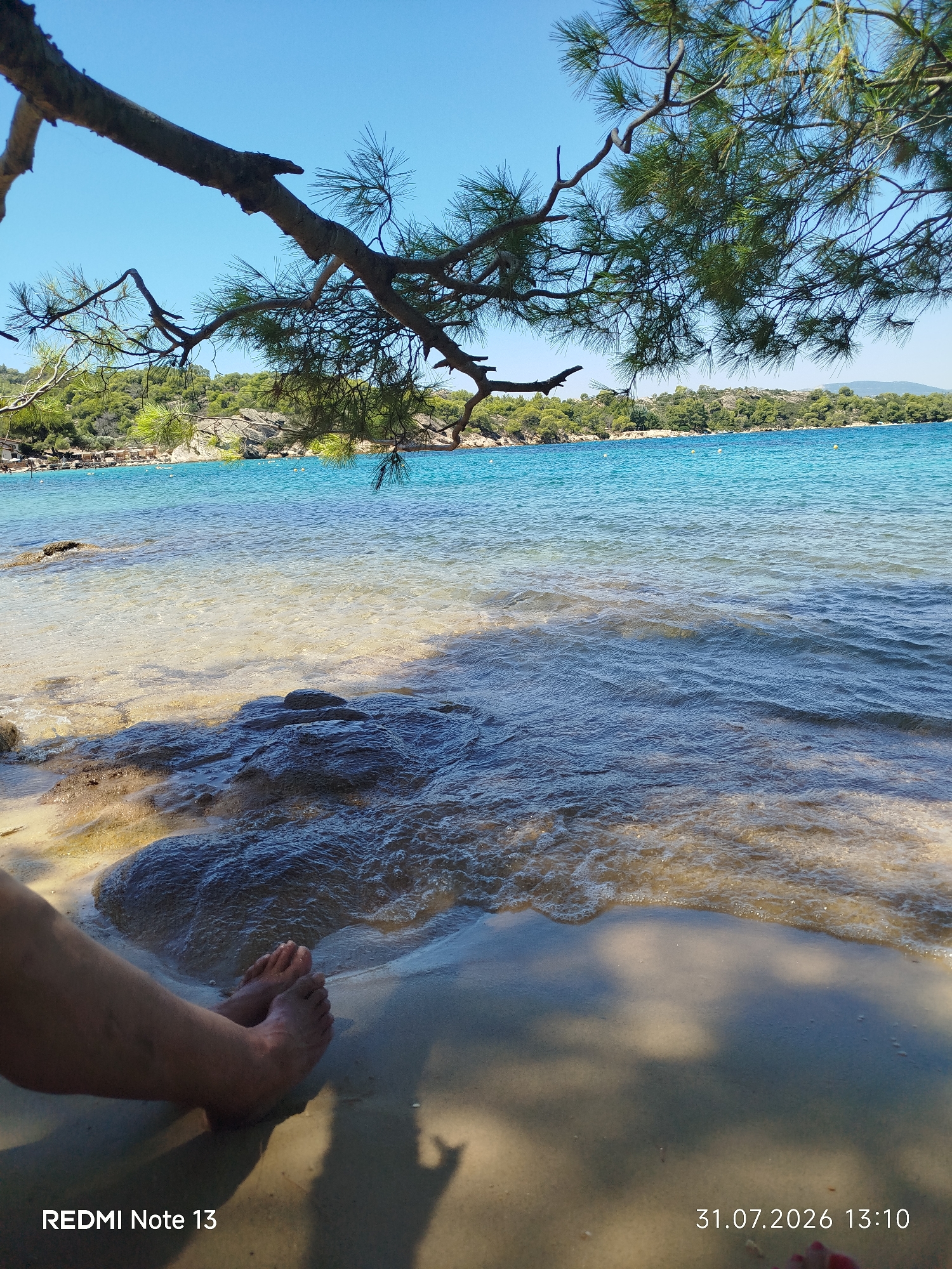 In Sithonia Municipal Unit, Greece, a serene beach scene unfolds under a clear blue sky. The foreground is dominated by the sandy shore and the gentle lapping of turquoise water. A person's bare feet are visible on the wet sand, partially submerged by the incoming tide. Dark, wet rocks are scattered near the shoreline. In the midground, the shallow water is crystal clear, allowing the sandy seabed and scattered stones to be seen. The background features a verdant coastline with dense green foliage and distant hills. Pine branches extend from the upper left, creating a natural frame for the scene. The water appears calm with gentle ripples. A few small yellow buoys are visible in the distance, marking a swimming area. The overall impression is one of peaceful seaside relaxation.