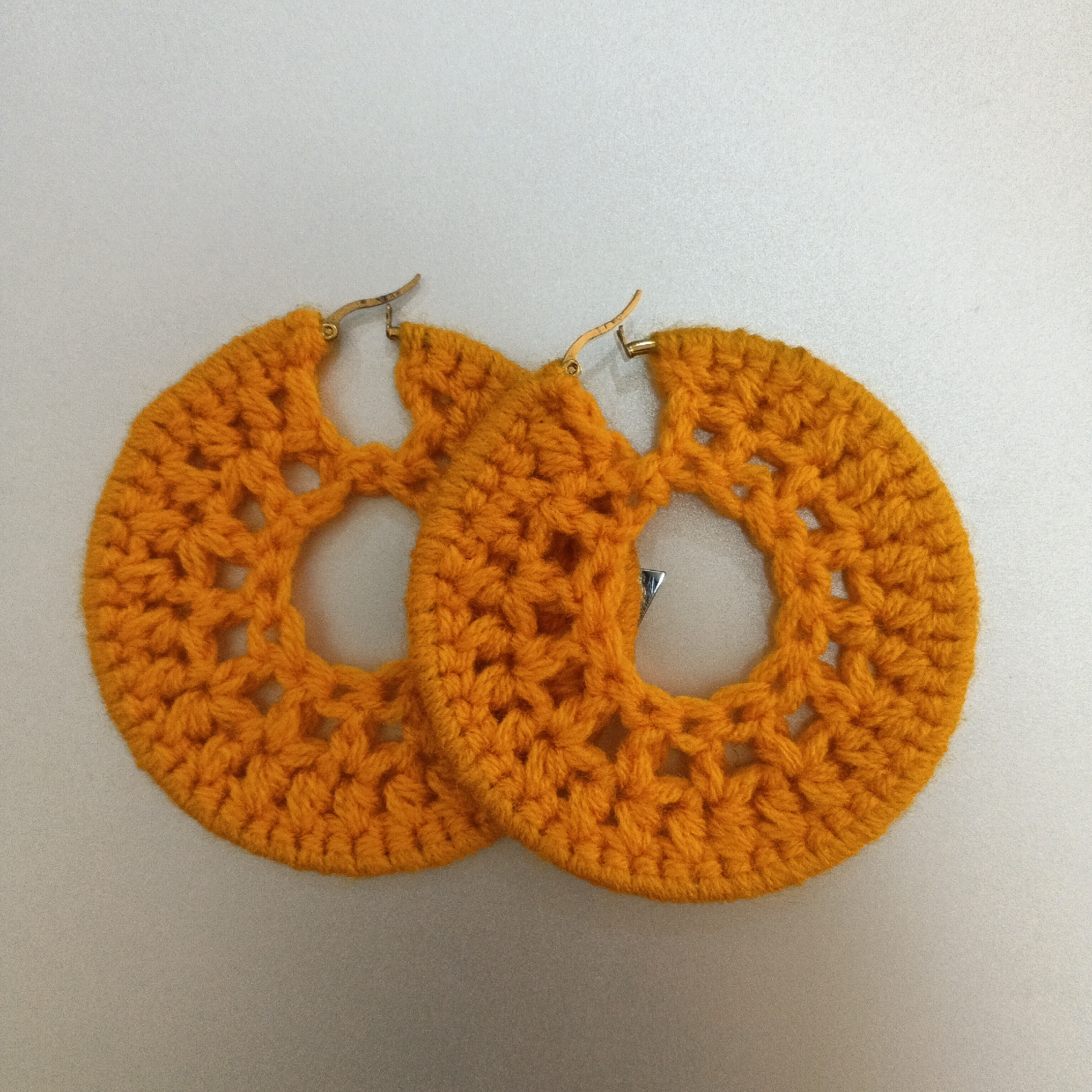 Two large, circular, orange crocheted hoop earrings are displayed against a plain, light-colored background. The earrings have an open, lacy pattern with geometric cutouts. Gold-colored earring hooks are attached to the top of each hoop. The earrings are positioned side-by-side, with the left earring slightly overlapping the right. The image is a top-down view. There is no discernible location in the city of Shaba, Nigeria.