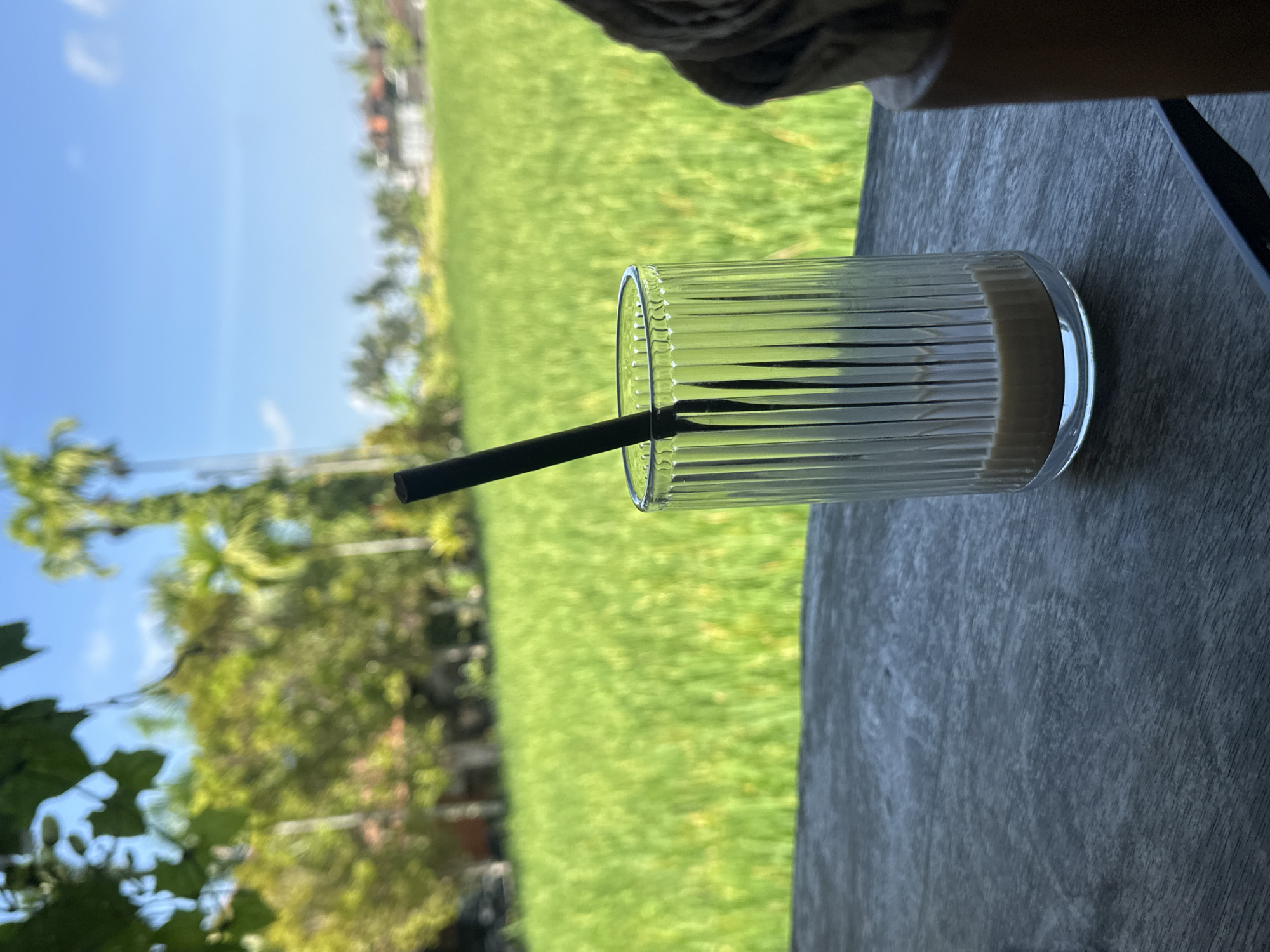 A glass of coffee with a straw sits on a wooden table, with a lush green rice paddy field and palm trees in the background under a clear blue sky. The scene is likely set in or around Ubud District, Indonesia, known for its picturesque rice terraces and tropical landscape.