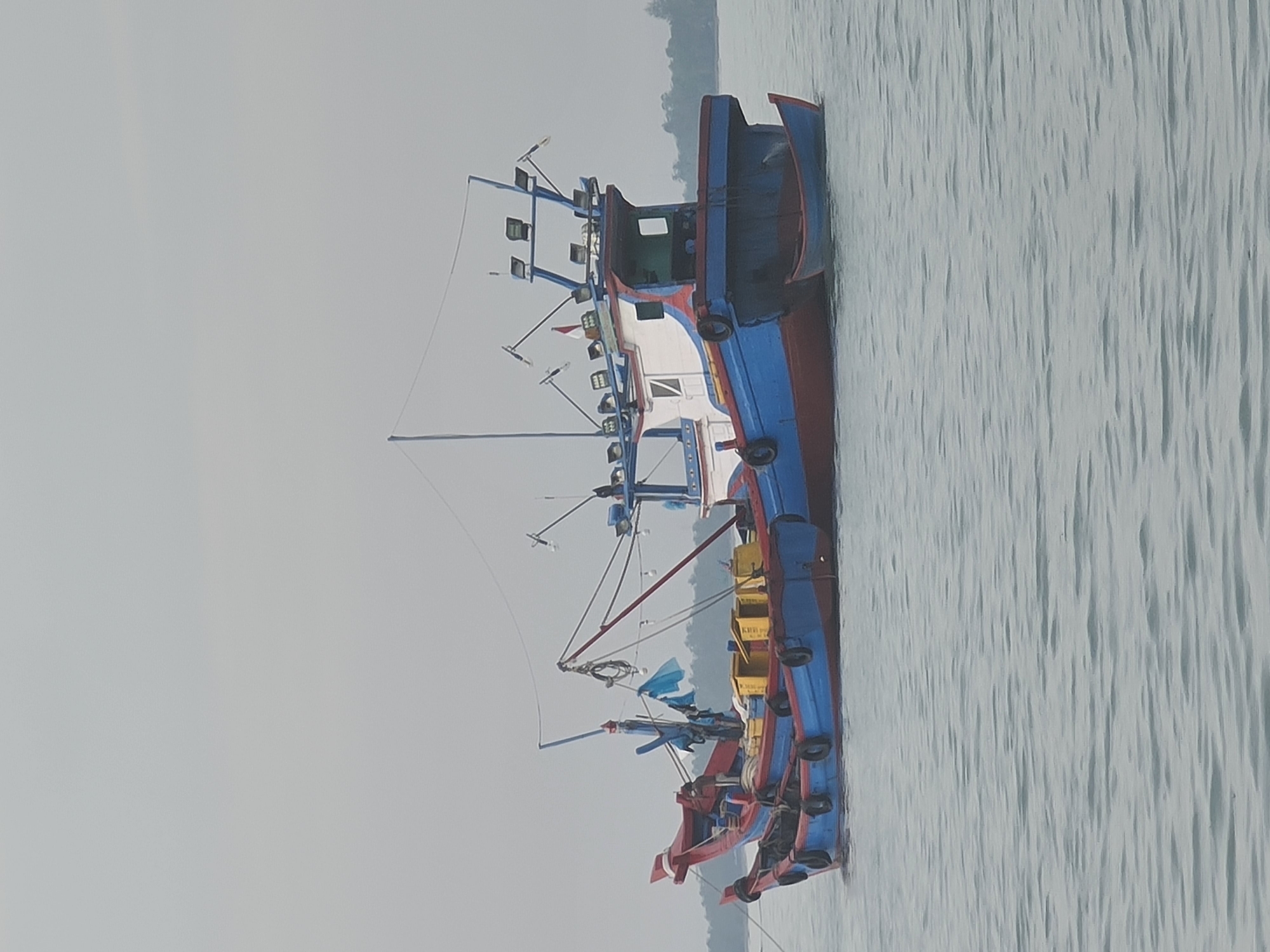 This image captures a fishing trawler docked on the calm waters near Lhokseumawe, Indonesia. The boat is painted in vibrant blue and red hues, with a white cabin structure equipped with multiple spotlights. Yellow containers are visible on the deck. A smaller, blue dinghy is tied to the side of the main vessel. The background shows a densely forested shoreline under an overcast sky. The water exhibits a rippled surface, suggesting minimal wave activity. There are no people or animals visible in the scene.