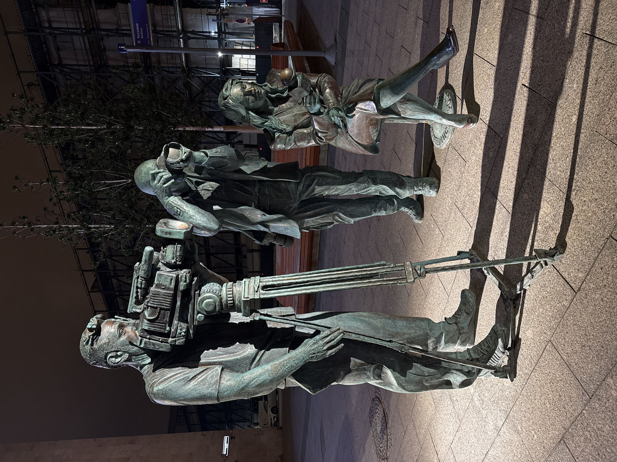 The image depicts a bronze sculpture of a film crew located in Moscow, Russia. The sculpture includes a cameraman with a large film camera, a sound technician with headphones, and an actress sitting on a director's chair. The scene is illuminated by artificial lighting at night. The location is identified by a sign in Russian, presumably indicating the "Российско" or "Rossiisko" film studio.