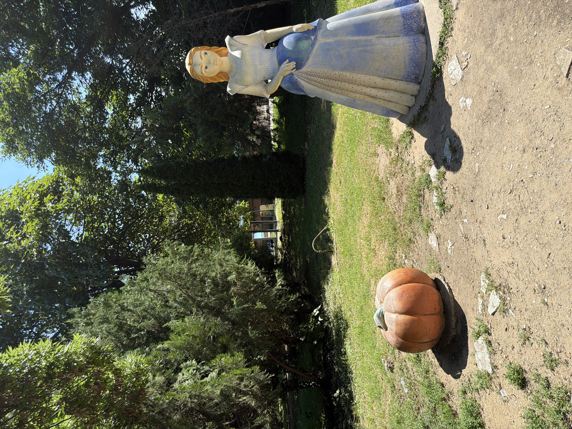 The image depicts an outdoor scene in the city of Pazardzhik, Bulgaria, featuring a statue of a woman in a blue and white dress standing on a dirt path next to a large pumpkin sculpture. The statue, which appears to be made of concrete or plaster, is positioned to the right of the frame and is facing left. The woman has blonde hair and blue eyes, and her dress is adorned with intricate patterns.
To the left of the statue is a round, orange pumpkin sculpture, also made of concrete or plaster. It rests on a circular stone base. The ground around the sculptures is a mix of dirt and patchy grass, with scattered stones and debris.
In the background, lush green trees and bushes form a dense canopy, suggesting a park or garden setting. Sunlight filters through the leaves, creating dappled shadows on the ground. Further in the background, beyond the trees, structures and possibly people are visible, indicating the presence of other park amenities or a nearby area. The overall ambiance is one of a sunny day in a public outdoor space.