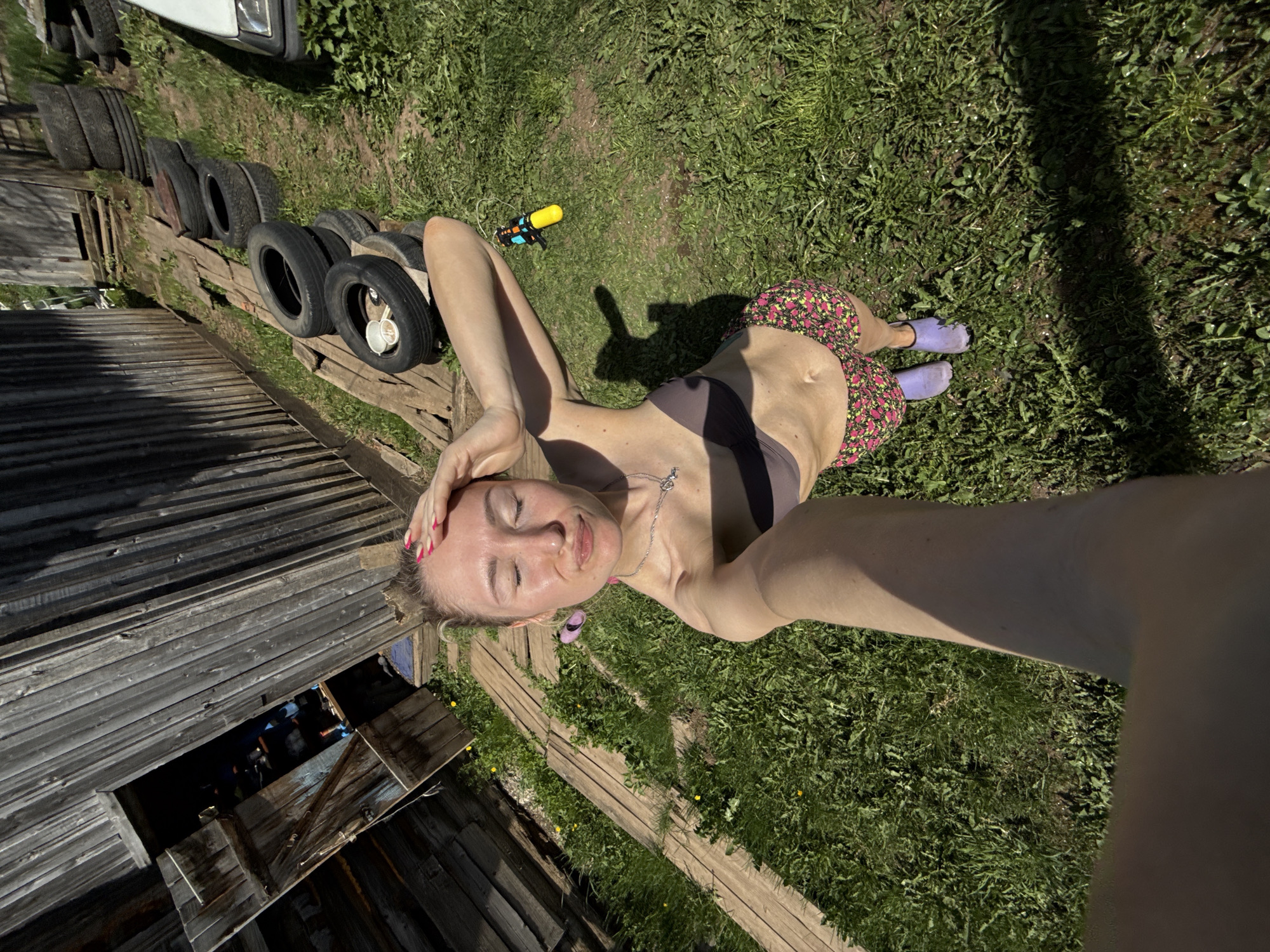 The image is a selfie taken outdoors, possibly in Nizhnyaya Chermoda, Russia, based on the context. A woman with blonde hair is the central figure, lying on a wooden structure or platform. She is wearing a dark-colored bikini top and floral-patterned shorts, along with purple footwear.
Behind the woman is a grassy area with a yellow and blue water gun nearby. To her left is a stack of old tires, followed by a weathered wooden building with an open door or window revealing a darker interior. The scene is bathed in sunlight, casting shadows on the grass and the woman's body.