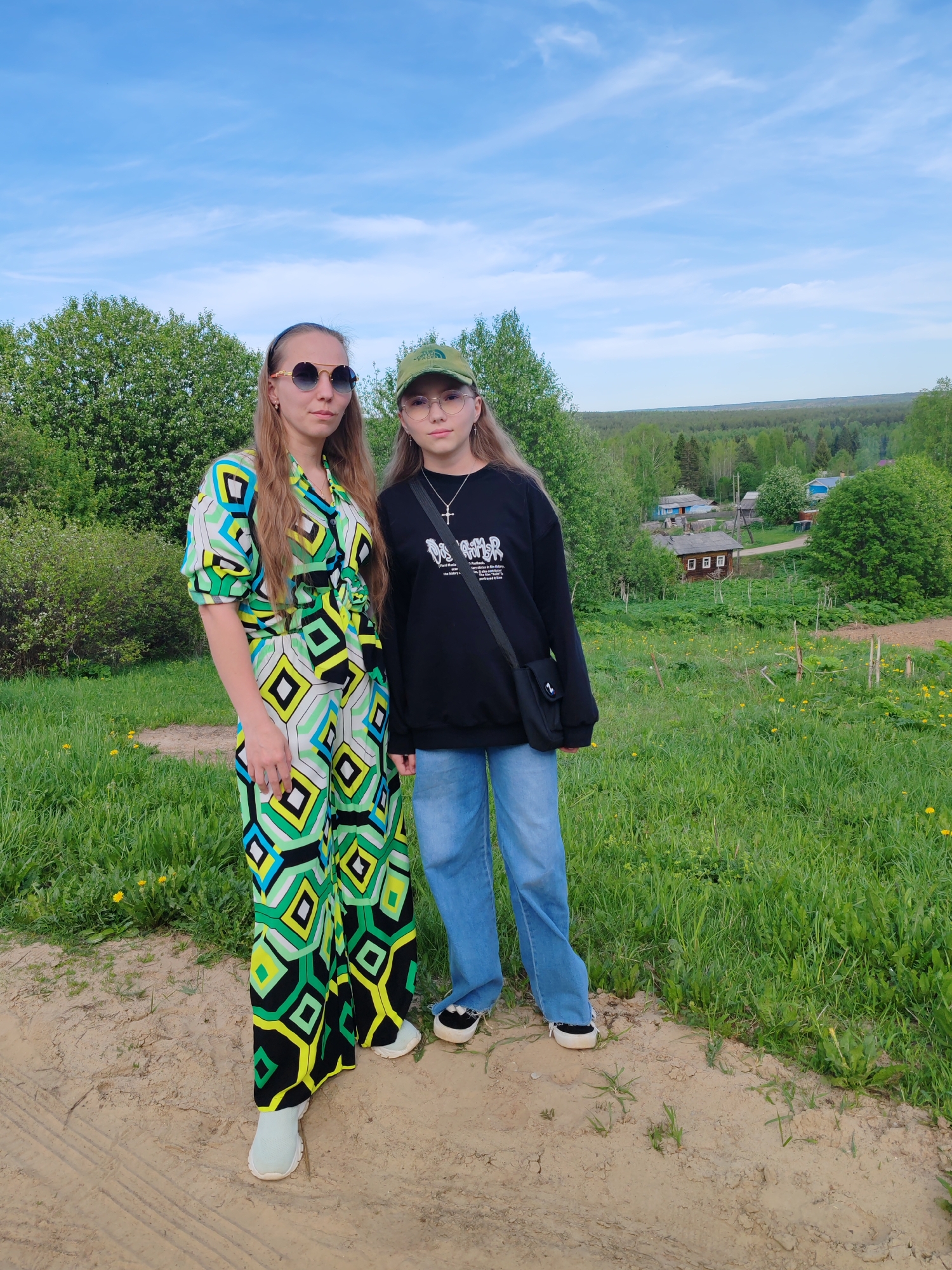 Two young women stand side-by-side on a dirt path in a grassy area. Behind them is a rural landscape with trees and houses, suggestive of a village on the outskirts of the city of Чухлэм, Russia. The woman on the left wears sunglasses, a long-sleeved, geometric-patterned jumpsuit in shades of green, black, and white, and light-colored shoes. The woman on the right wears glasses, a green baseball cap, a black sweatshirt with white writing, light blue jeans, and black and white sneakers. She has a black cross necklace. The two stand close together, looking toward the camera. The sky above is light blue with some clouds. The background features green trees and houses, indicative of a rural or semi-rural setting.